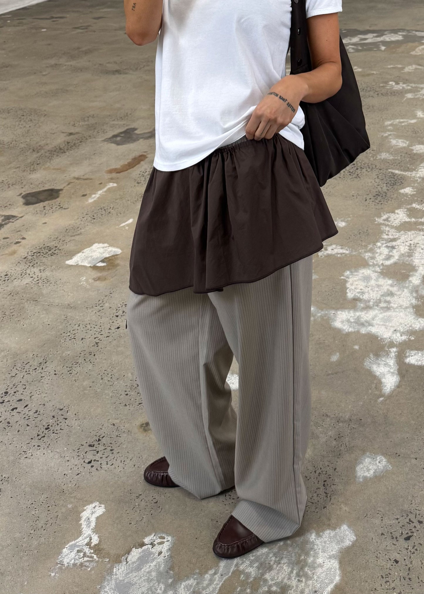 A person stands on a textured concrete floor, wearing a white organic cotton t-shirt, brown skirt over gray pinstriped wide-leg pants, brown shoes, a dark bag, and the Napkin Belt - Chocolate. Their face is not visible.