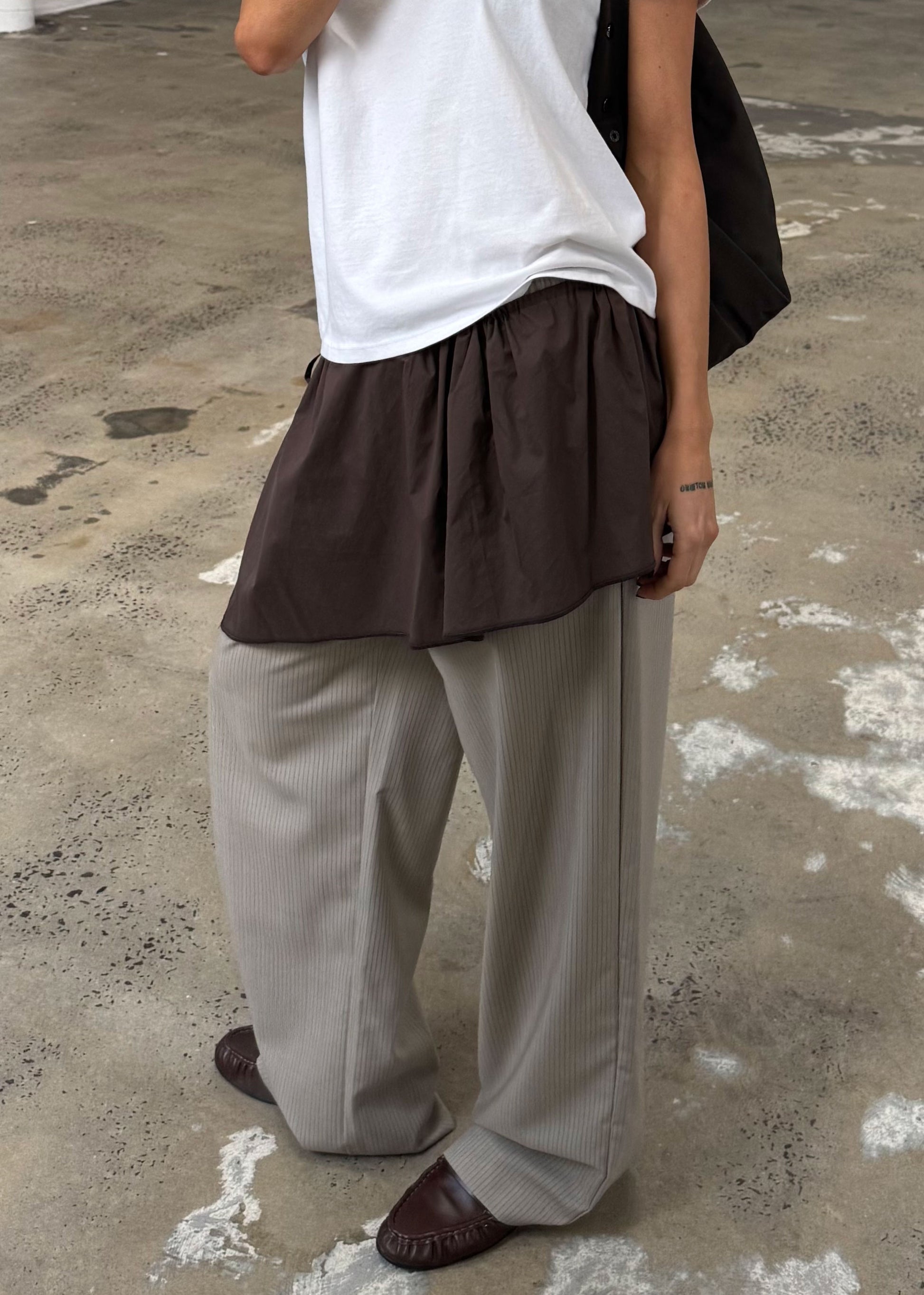 A person indoors wears a white organic cotton tee, a brown skirt over gray wide-leg pants, brown shoes, and carries a black bag. Their look is accented by the Napkin Belt - Chocolate. The floor is worn concrete with paint marks.