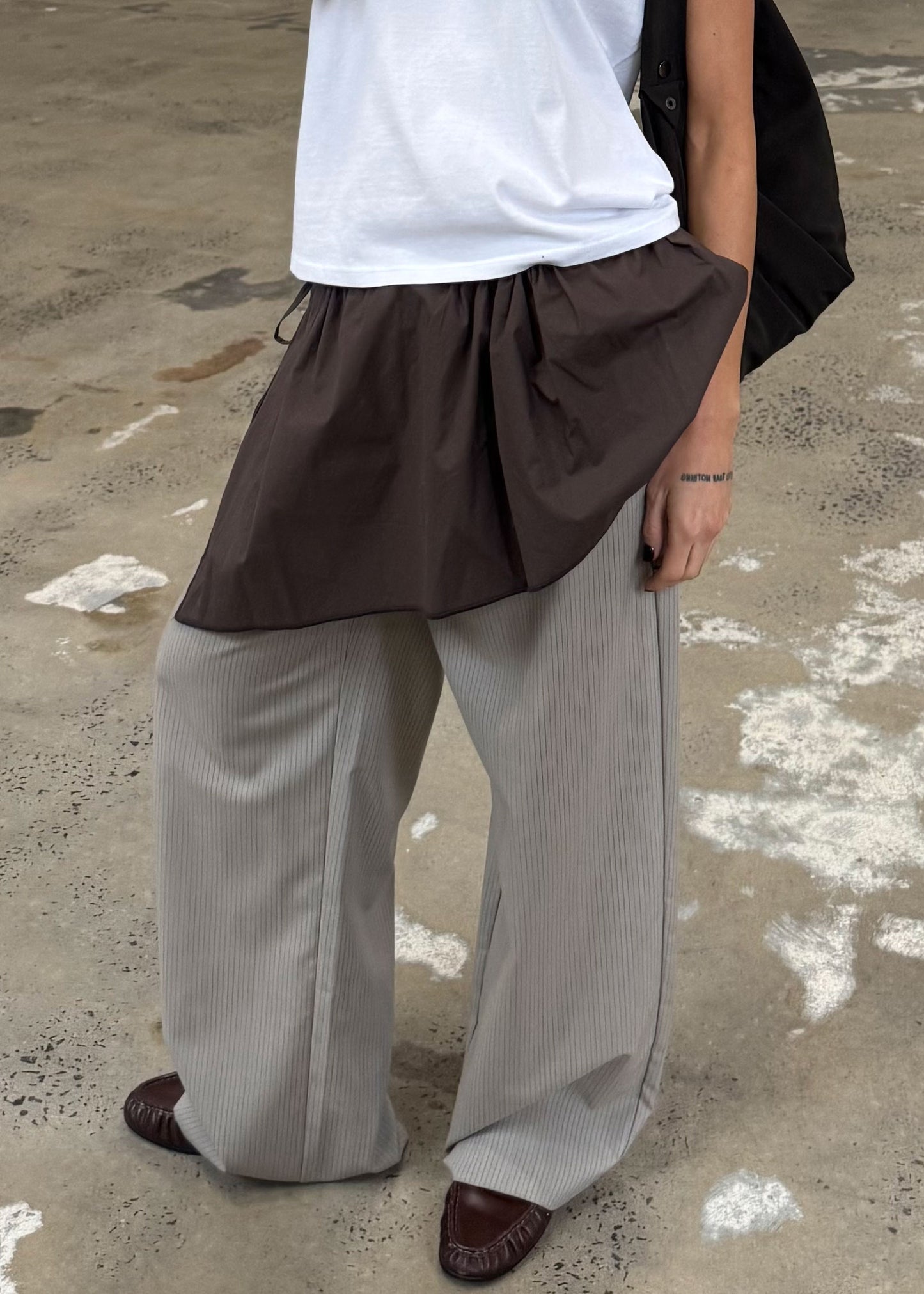 A person stands on a concrete floor in a white t-shirt, brown skirt over wide gray pinstriped pants, brown shoes, and wears the Napkin Belt - Chocolate by Deiji Studios. A small tattoo is visible near their right wrist.