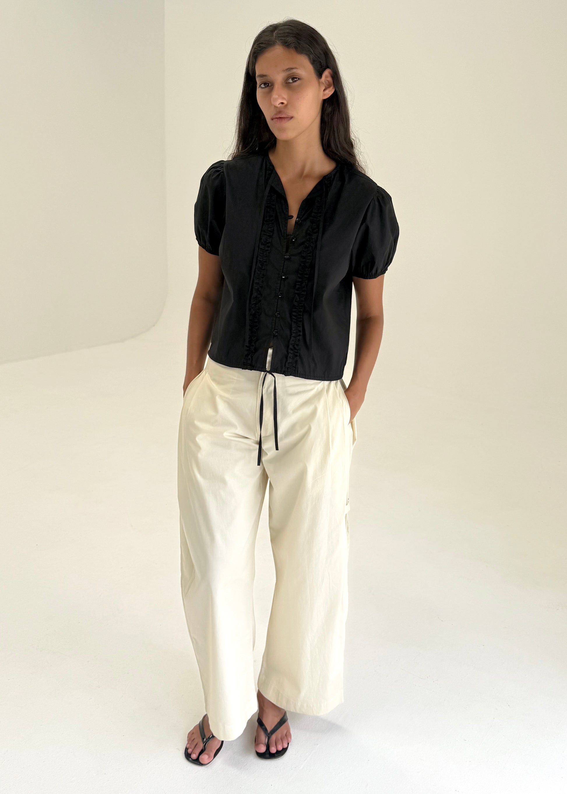 A woman with long brown hair stands in a minimal white space wearing Deiji Studios’ Double Frill Top in black, cream wide-leg drawstring pants, and black sandals, her hands tucked into her pockets.