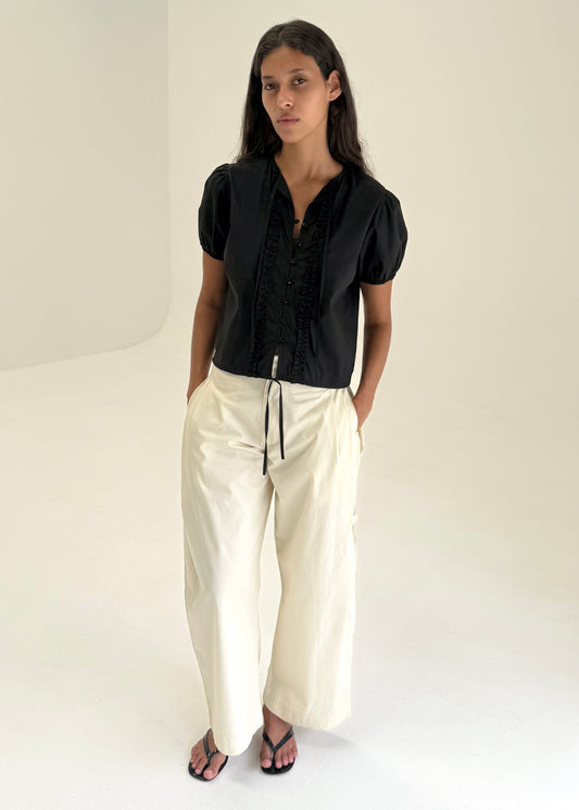 A woman with long brown hair stands in a minimal white space wearing Deiji Studios’ Double Frill Top in black, cream wide-leg drawstring pants, and black sandals, her hands tucked into her pockets.