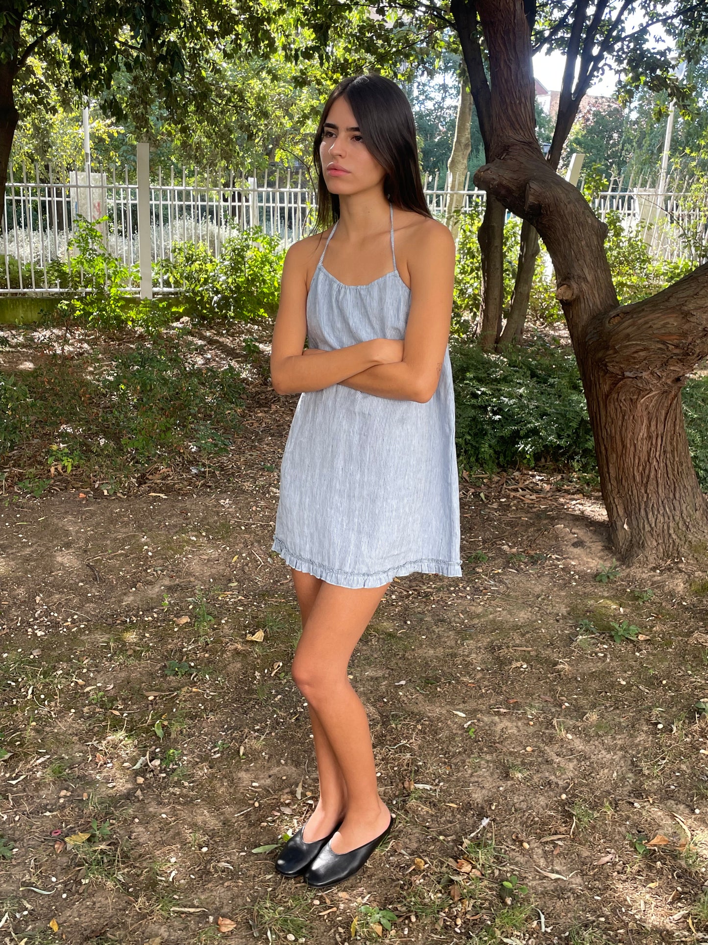 A young woman in a blue stripe Flounce Halter Dress and black shoes stands outdoors on dirt with her arms crossed, gazing to the side. Lush trees and greenery fill the background.