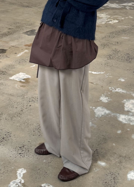 A person wearing loose beige organic cotton pants, a brown layered shirt, a dark blue sweater, and the Napkin Belt - Chocolate stands on a concrete floor with white patches. The image is cropped above the shoulders, so their face is not visible.