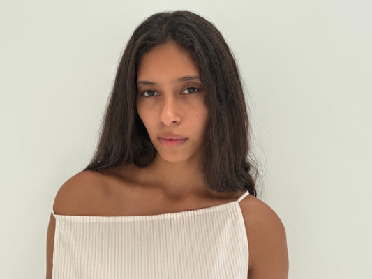 A young woman with long dark hair wears the Deiji Studios Simple Top in Dusty Stripe, made from organic cotton poplin, standing before a plain light background and looking calmly at the camera.