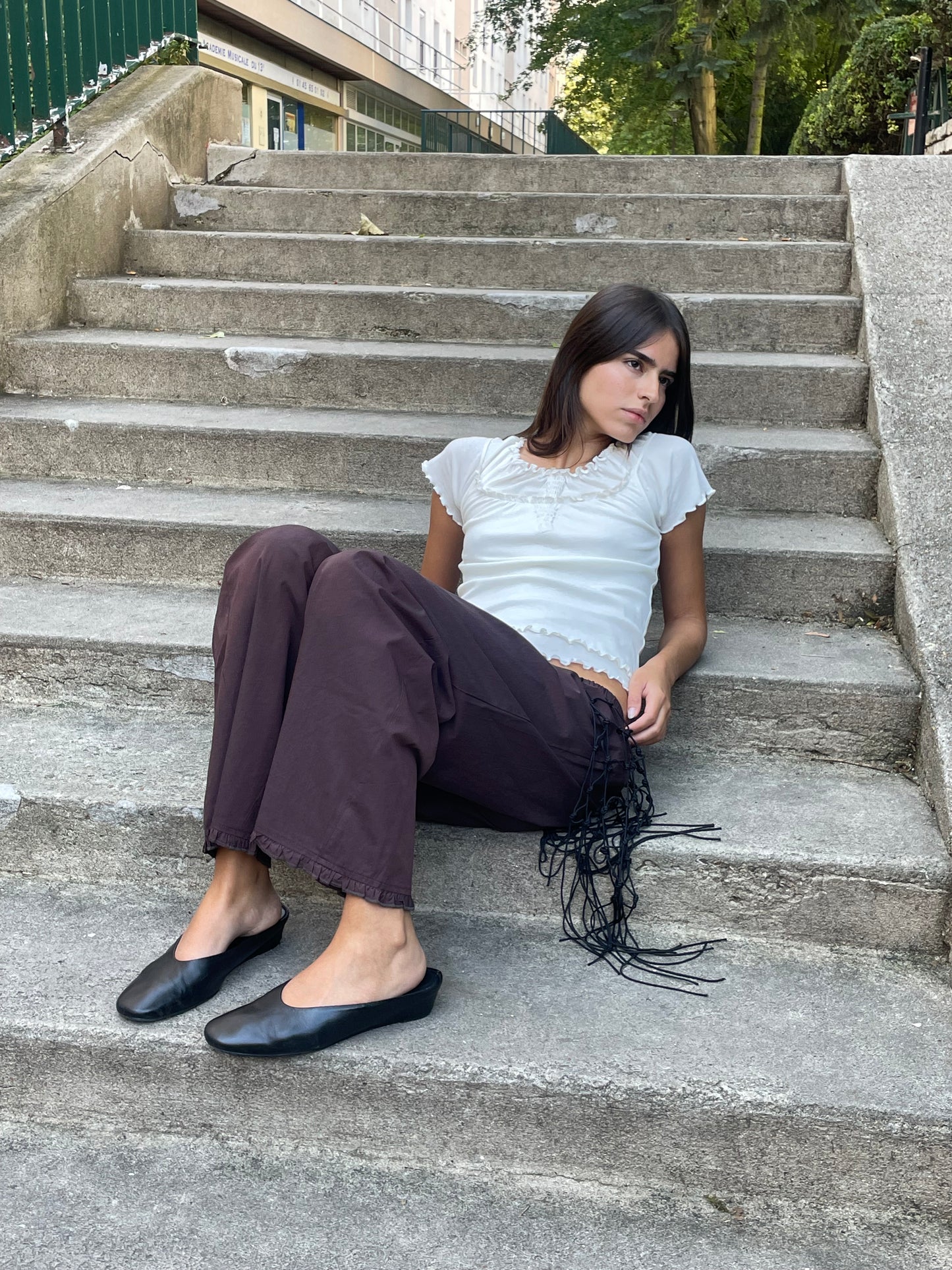 A young woman with long dark hair sits on outdoor concrete steps, wearing the Flounce Pant - Umber, a loose brown pair with fringe detail made from organic cotton, paired with a white short-sleeve top and black flats. She gazes calmly to her right.