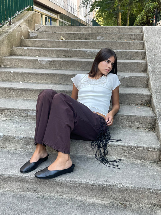 A young woman with long dark hair sits on outdoor concrete steps, wearing the Flounce Pant - Umber, a loose brown pair with fringe detail made from organic cotton, paired with a white short-sleeve top and black flats. She gazes calmly to her right.