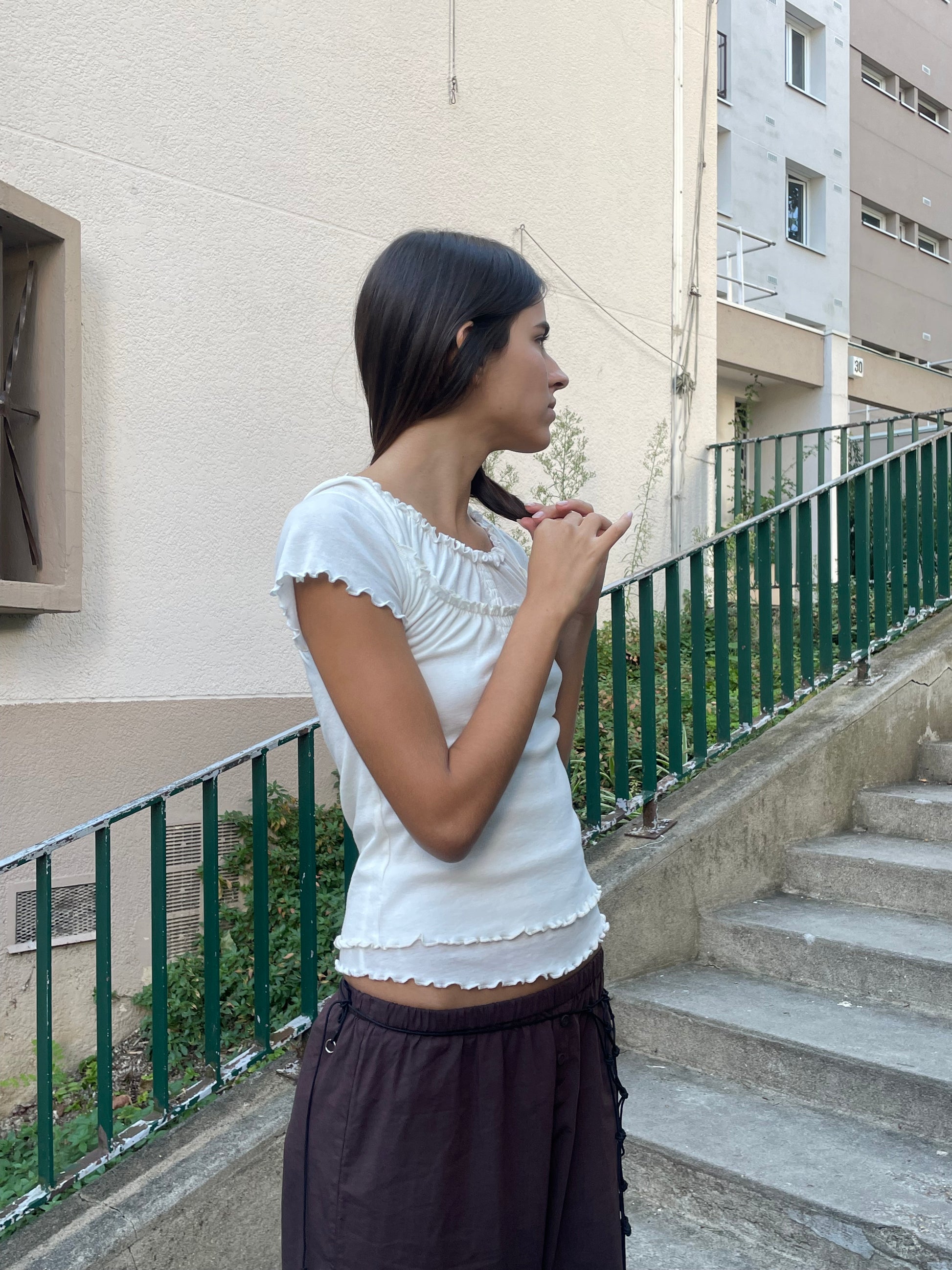A woman with long dark hair stands outdoors by a green railing and concrete steps, wearing Deiji Studios' Flounce Tee in Cream with ruffled edges and loose brown pants. She looks to the side, hands clasped together.