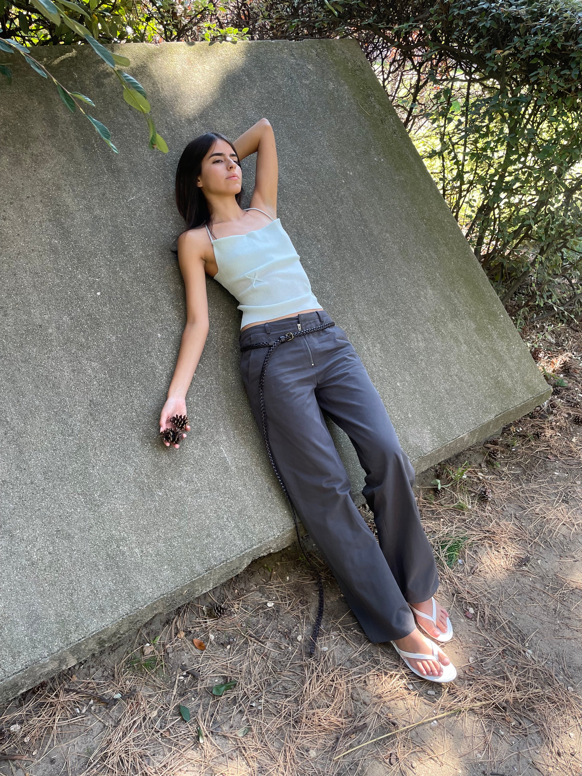 A young woman in the Fine Knot Tank - Ice Blue by Deiji Studios and dark pants reclines on a slanted concrete surface outdoors, surrounded by greenery and pine needles, holding a pinecone.