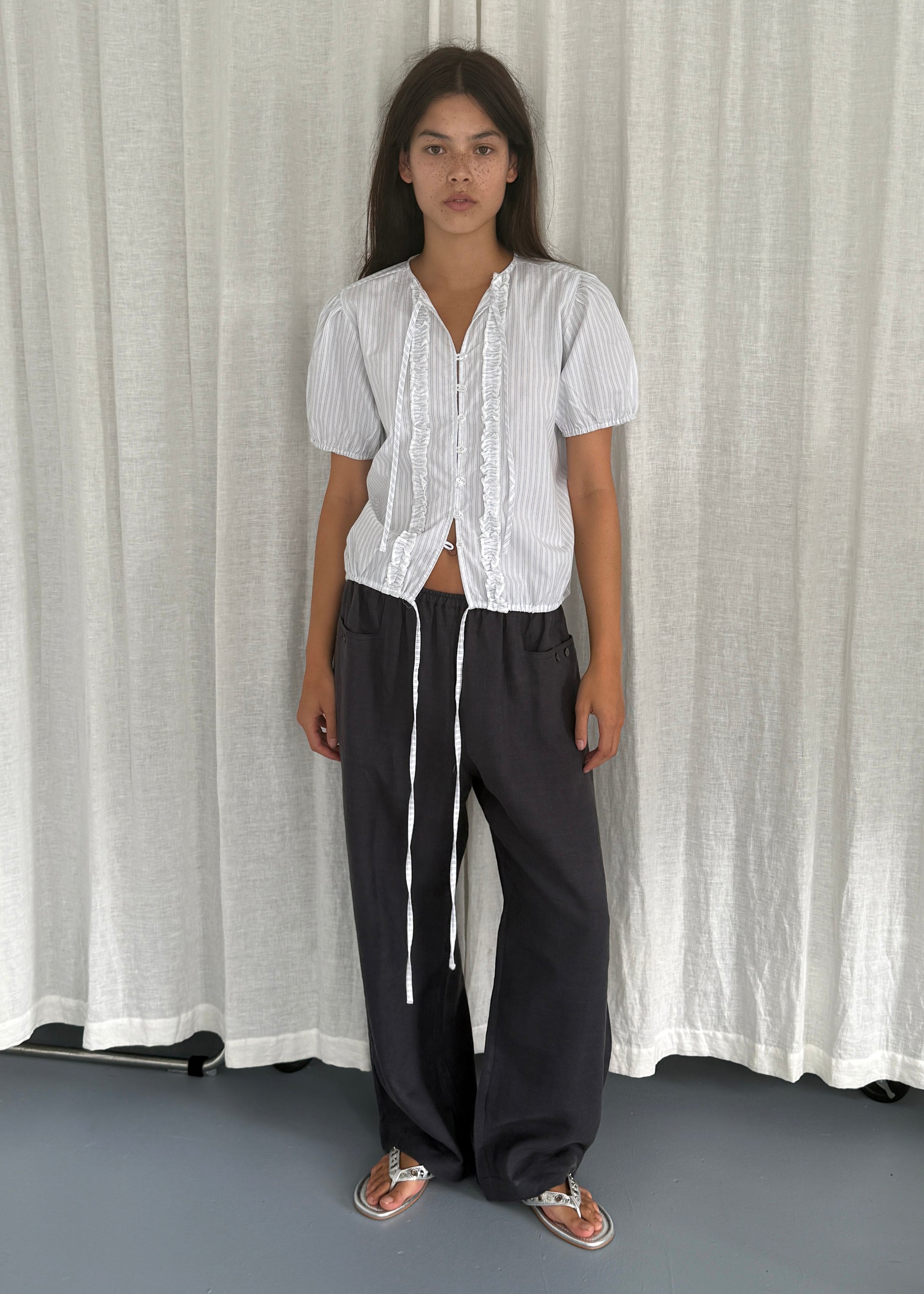 A woman stands in front of white curtains wearing the Double Frill Top - Sage Stripe by Deiji Studios, loose dark drawstring pants, and silver sandals. She faces the camera with a neutral expression.