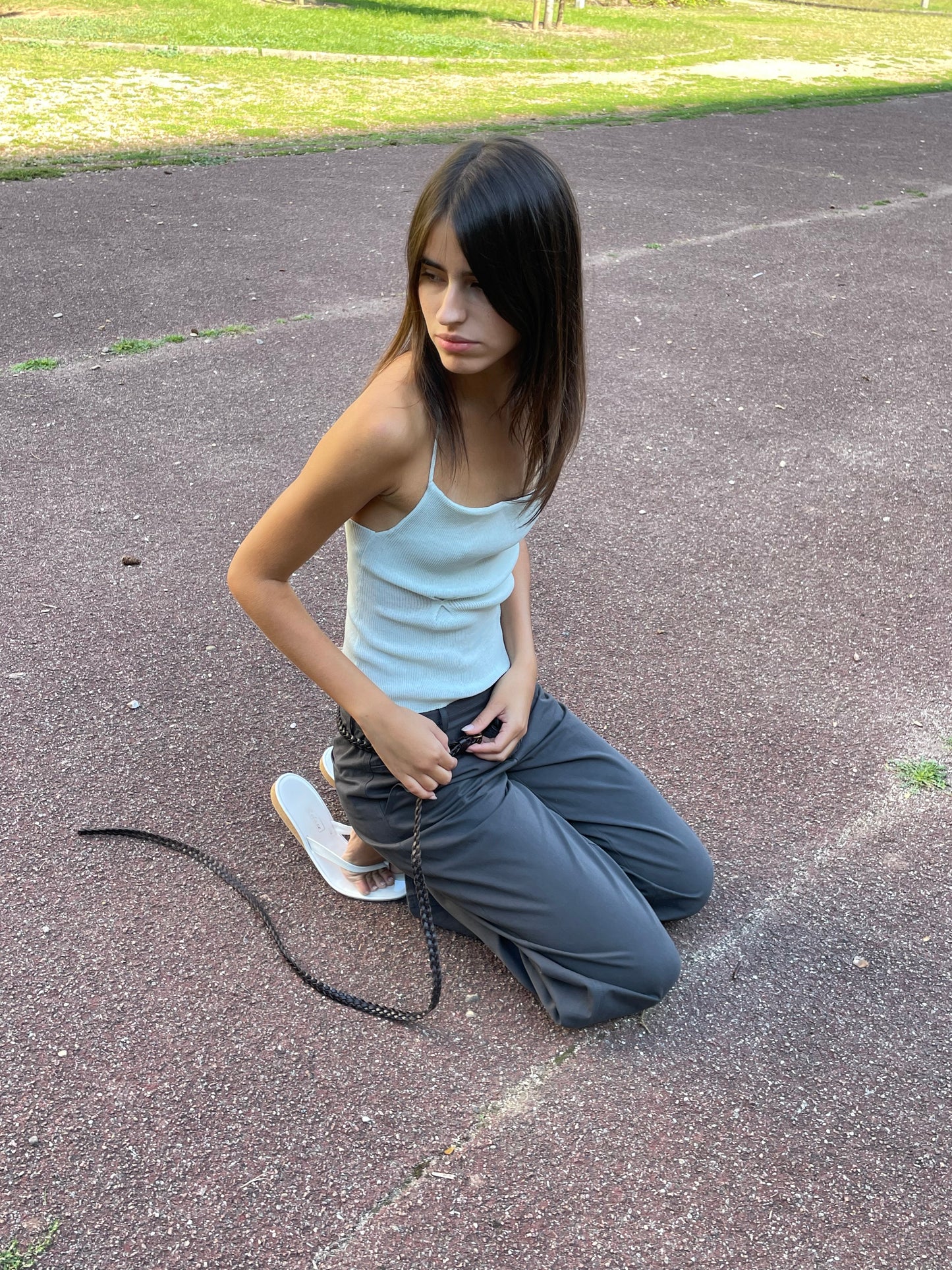 A young girl with long dark hair kneels outdoors in a light blue tank top and Deiji Studios Zip Pant - Charcoal, paired with white sandals. She holds a black rope, looking left, with grass visible in the background.