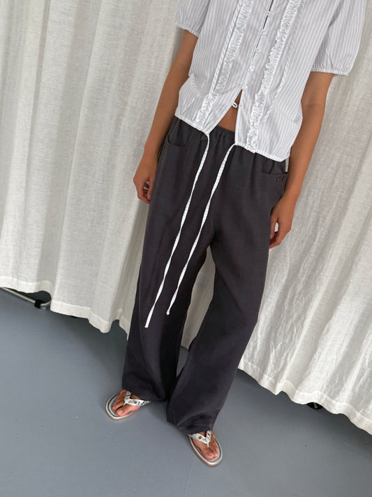 A person stands in front of white curtains wearing the Double Frill Top - Sage Stripe from Deiji Studios, featuring short sleeves, ties, and partially unbuttoned details, paired with loose dark gray pants and silver sandals. Their face is out of frame.