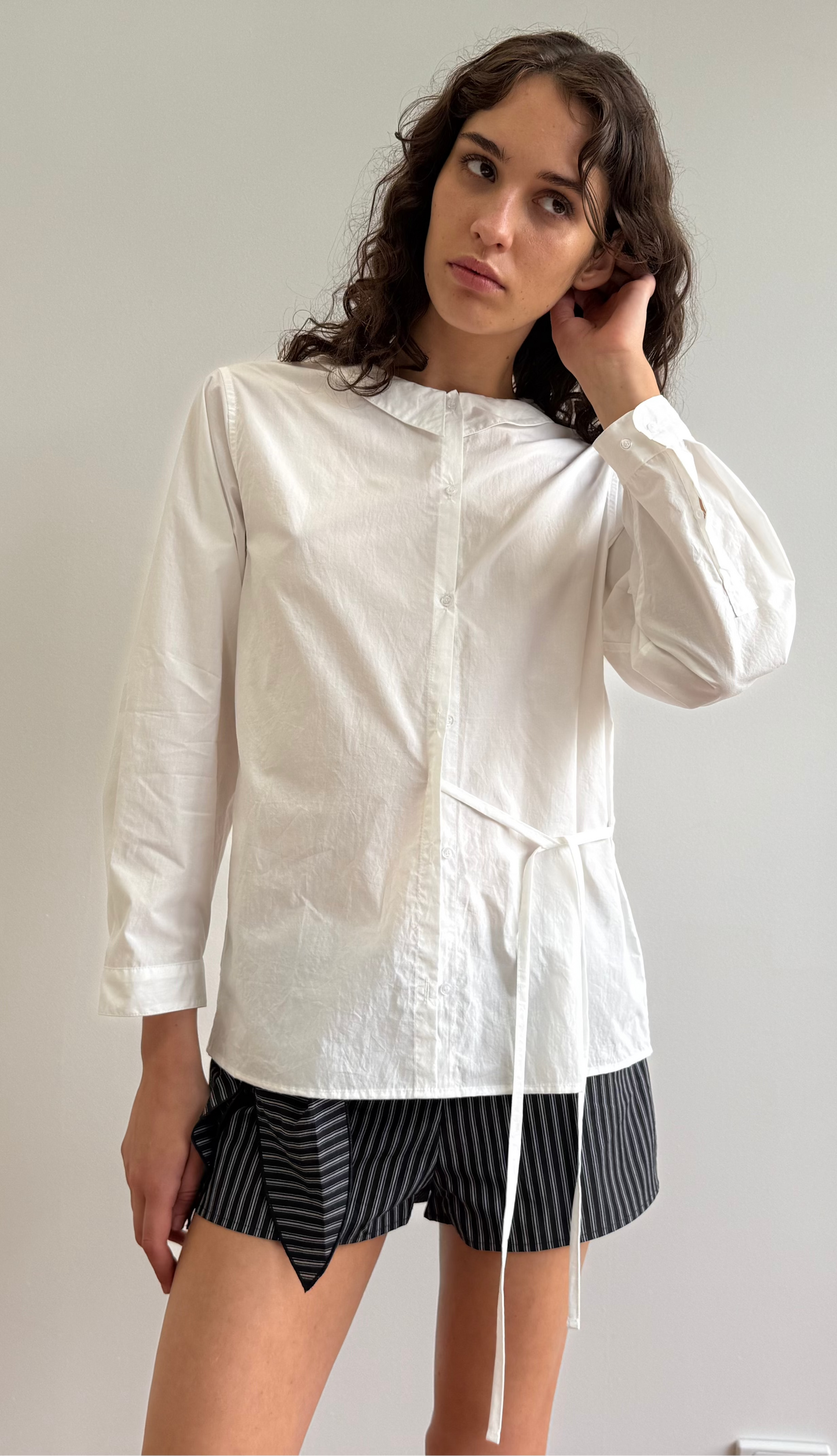 A person with long, wavy brown hair stands against a plain background, wearing the Left Shirt - White with an adjustable side tie and black-and-gray striped shorts, looking slightly to the side while touching their hair.
