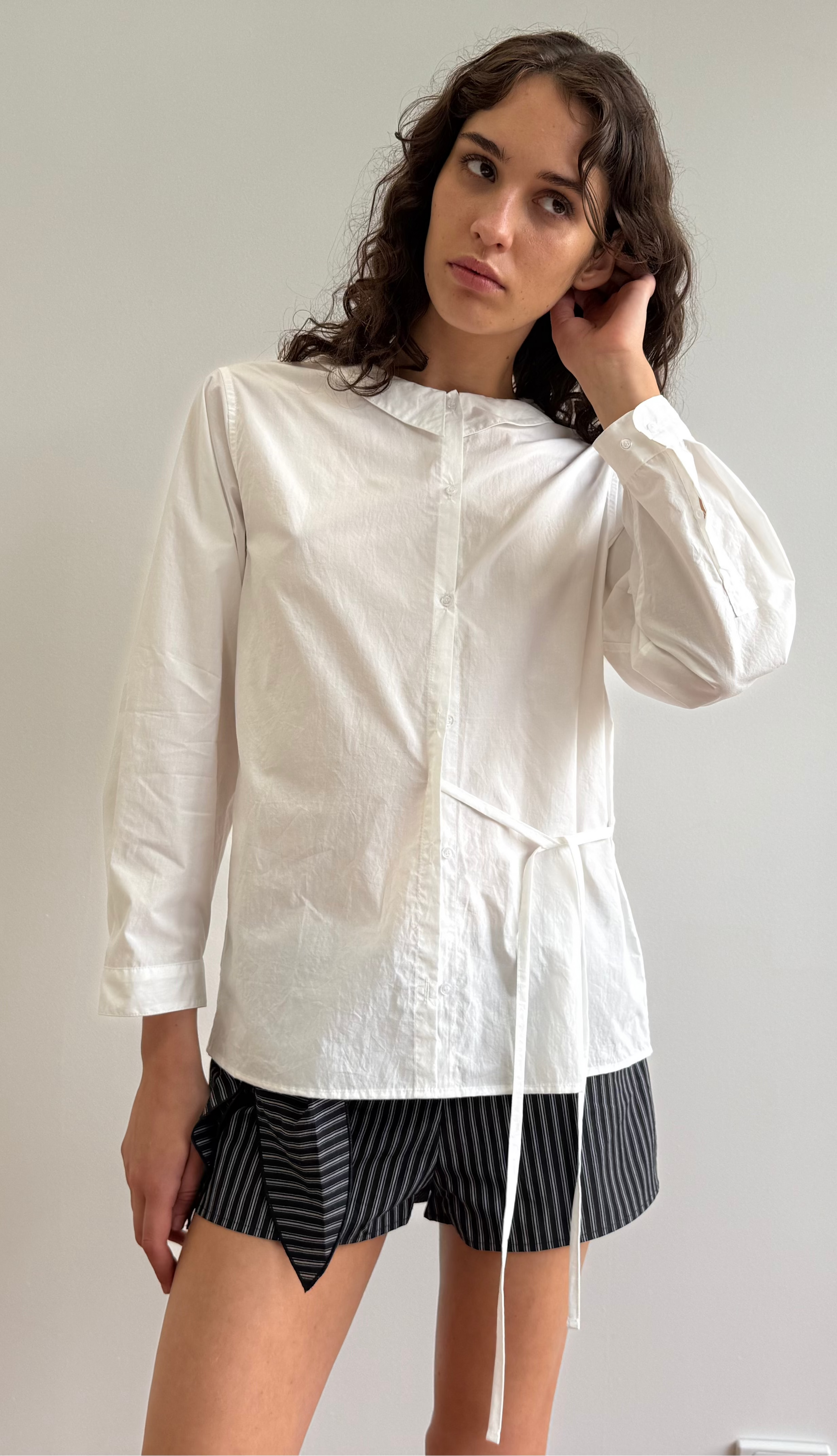A person with long, wavy brown hair stands against a plain background, wearing the Left Shirt - White with an adjustable side tie and black-and-gray striped shorts, looking slightly to the side while touching their hair.