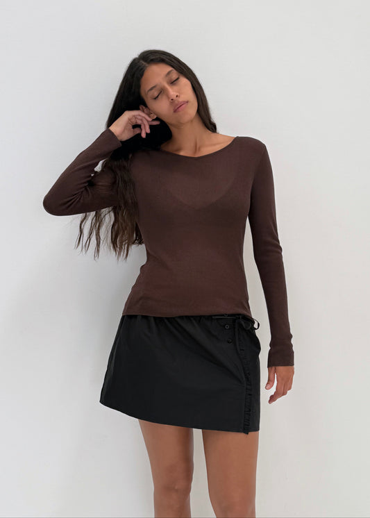 A woman with long brown hair, wearing the Knitted Top - Dark Brown and a black mini skirt, stands against a plain white background with her eyes closed and one hand in her hair.