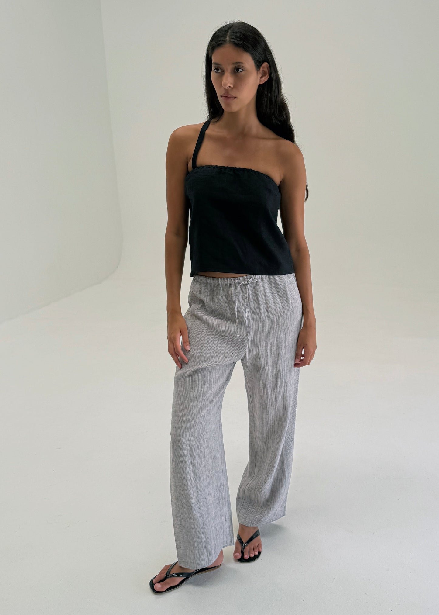 A woman with long dark hair stands in a minimal studio wearing a black strapless top, Double Waist Linen Pant in Haze Blue, and black sandals. She looks slightly to the side with a neutral expression.