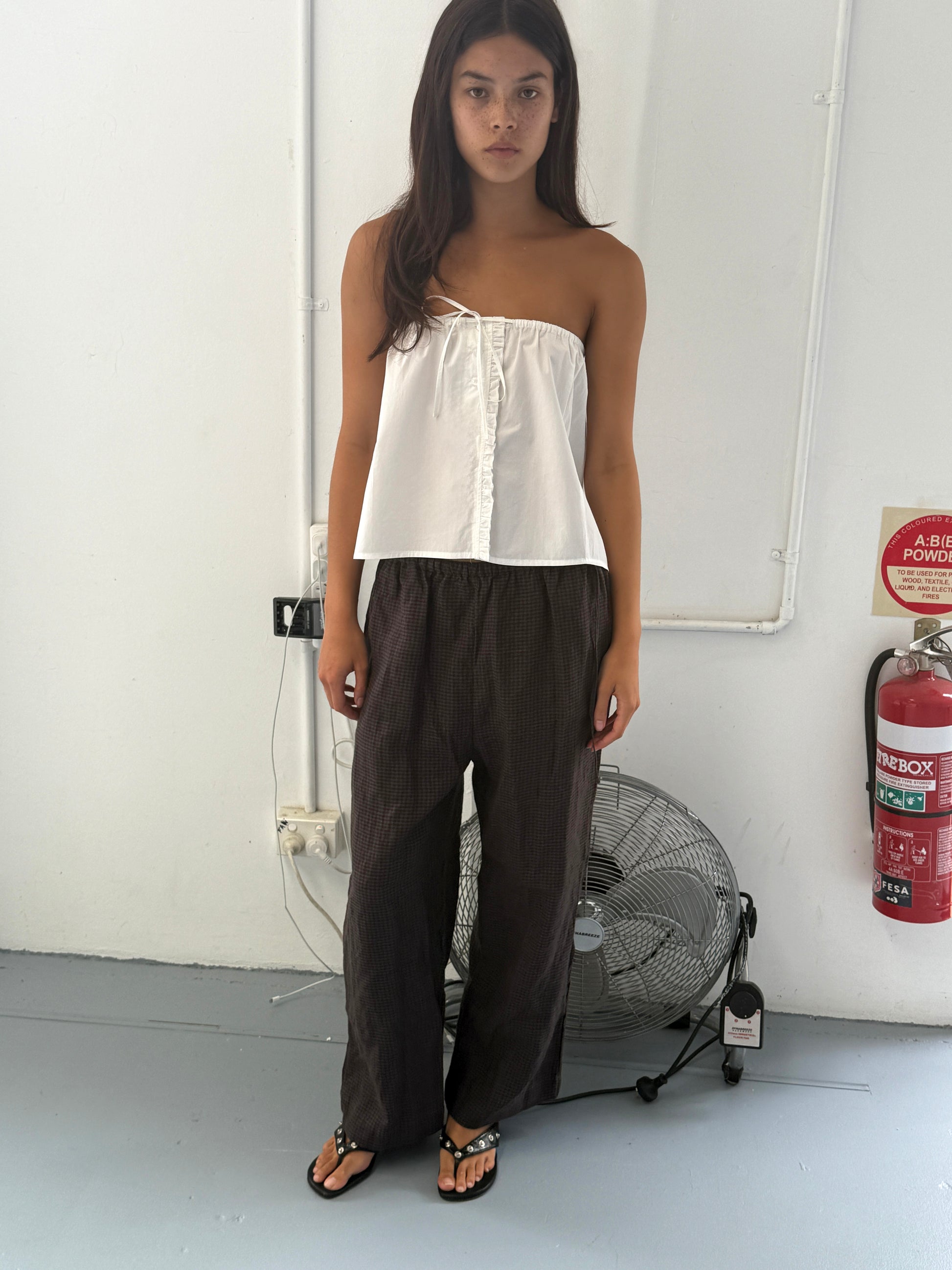 A woman with long dark hair stands indoors wearing a white strapless top, the Ruffle Mini Skirt - White, and sandals. Behind her are a large fan, a fire extinguisher, and exposed wall pipes.