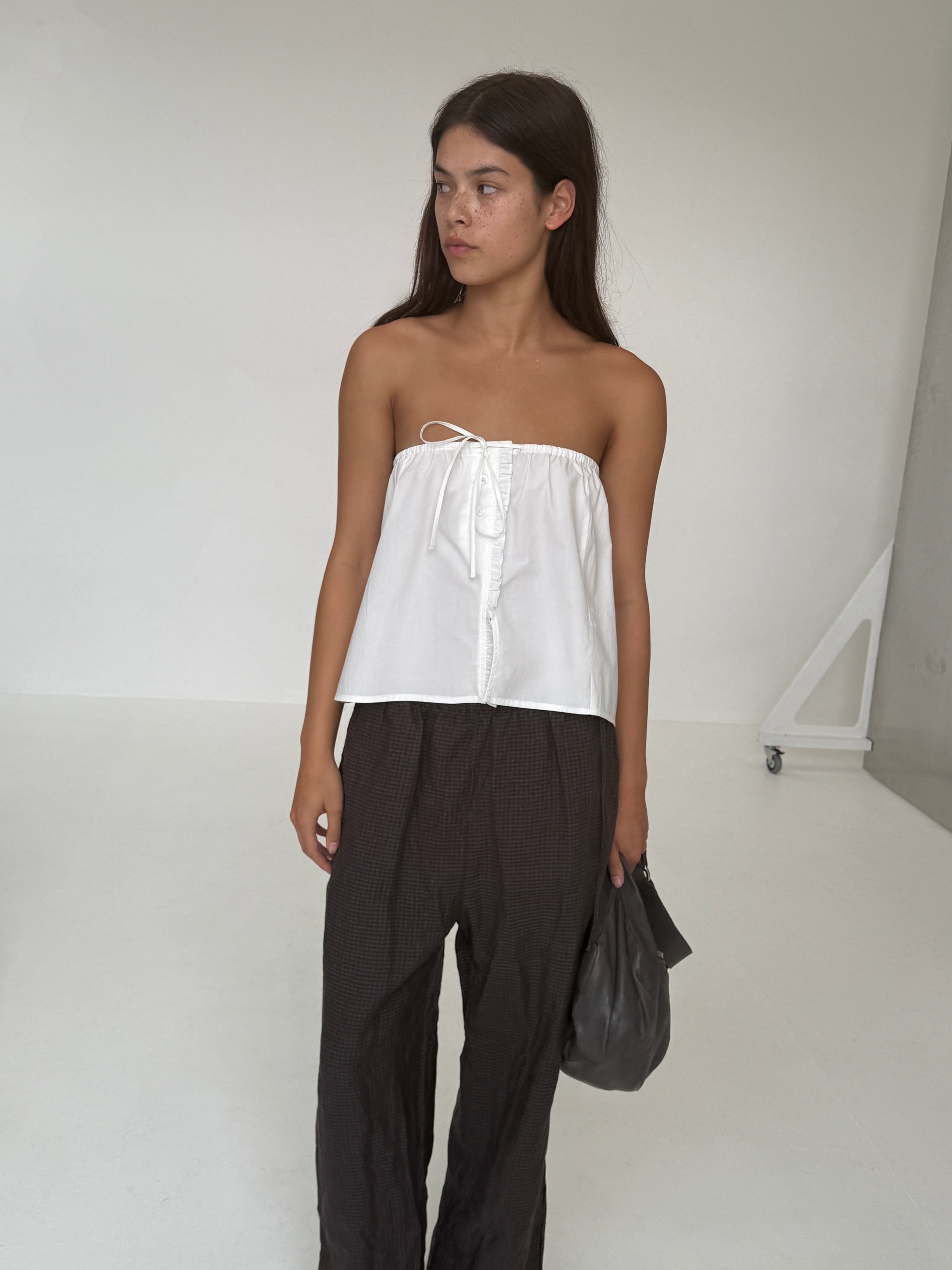 A young woman with long dark hair stands in a minimalist white room, wearing a Deiji Studios Ruffle Mini Skirt in white and loose dark pants. She holds a dark bag in her left hand and looks to the side with a neutral expression.
