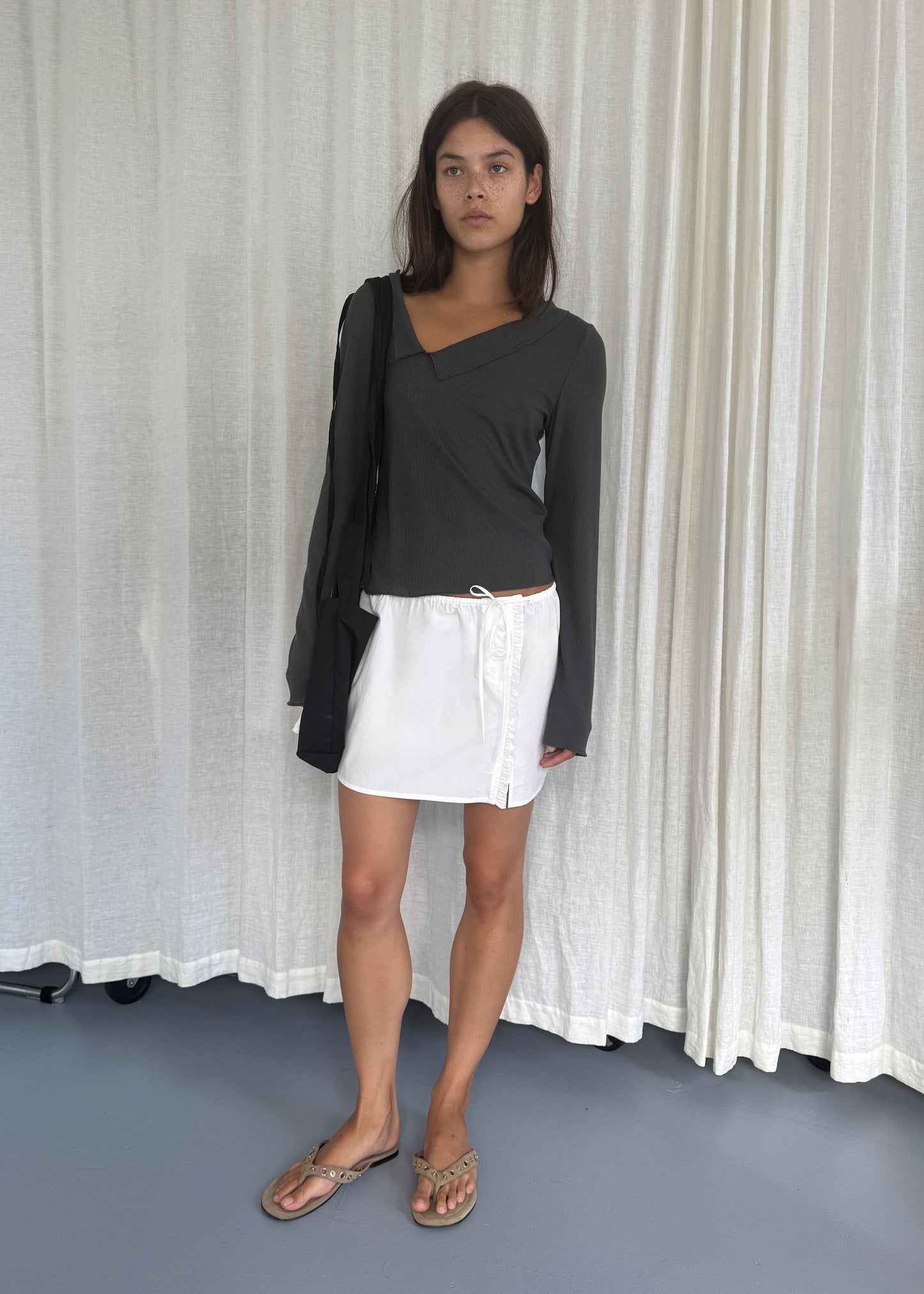 A young woman stands indoors before white curtains, wearing a dark long-sleeve top, the Ruffle Mini Skirt - White by Deiji Studios, brown sandals, and carrying a black bag over her shoulder.