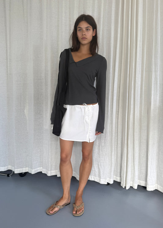 A woman with straight brown hair stands before white curtains, wearing a Deiji Studios Cross Collar Top in Night Blue, a short white skirt, flip-flops, and carrying a black bag over one shoulder.