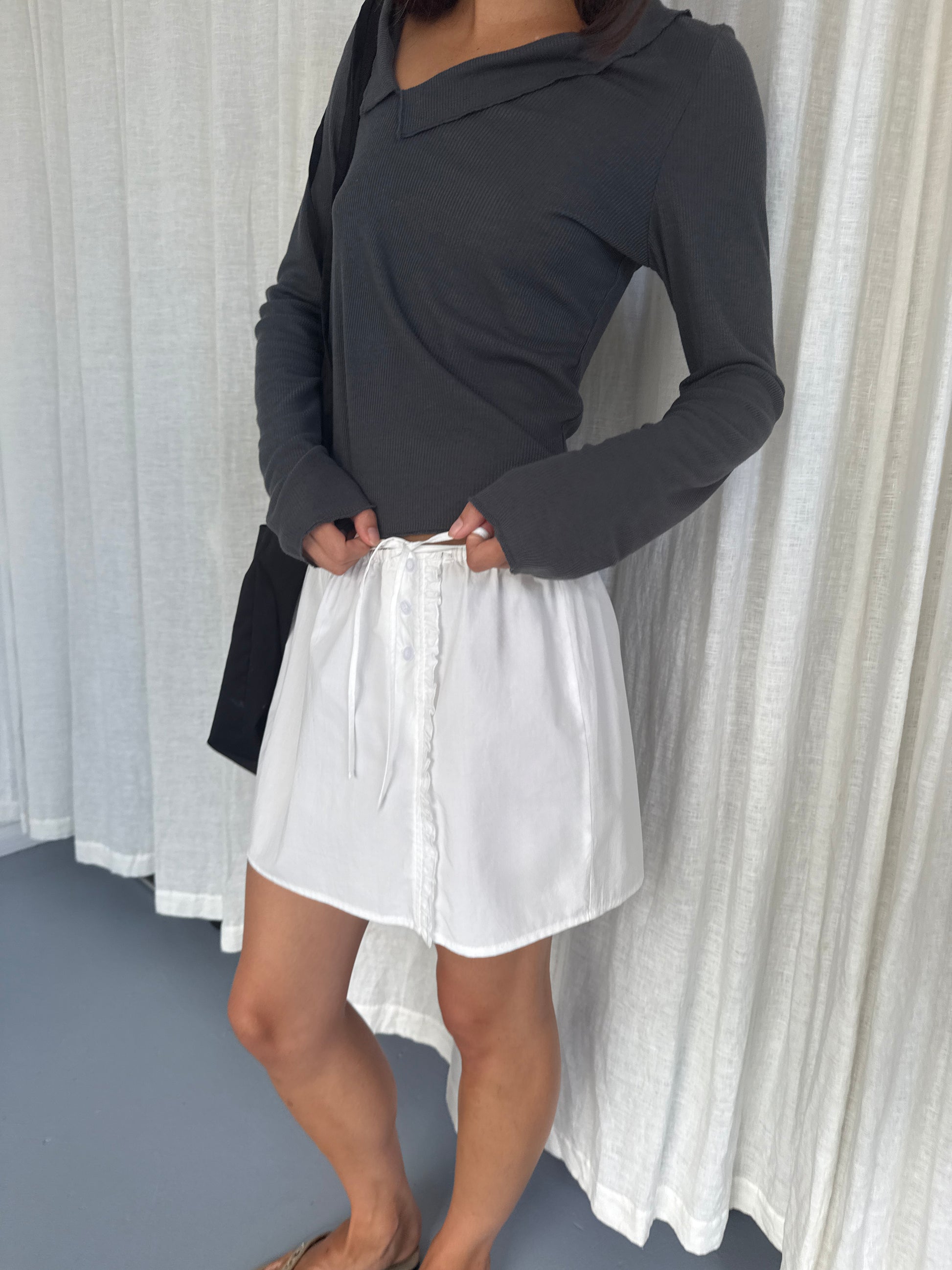 A person stands indoors wearing the Cross Collar Top in Night Blue, a long-sleeved sheer fine knit, paired with a white buttoned skirt and a black shoulder bag. White curtains hang in the background.