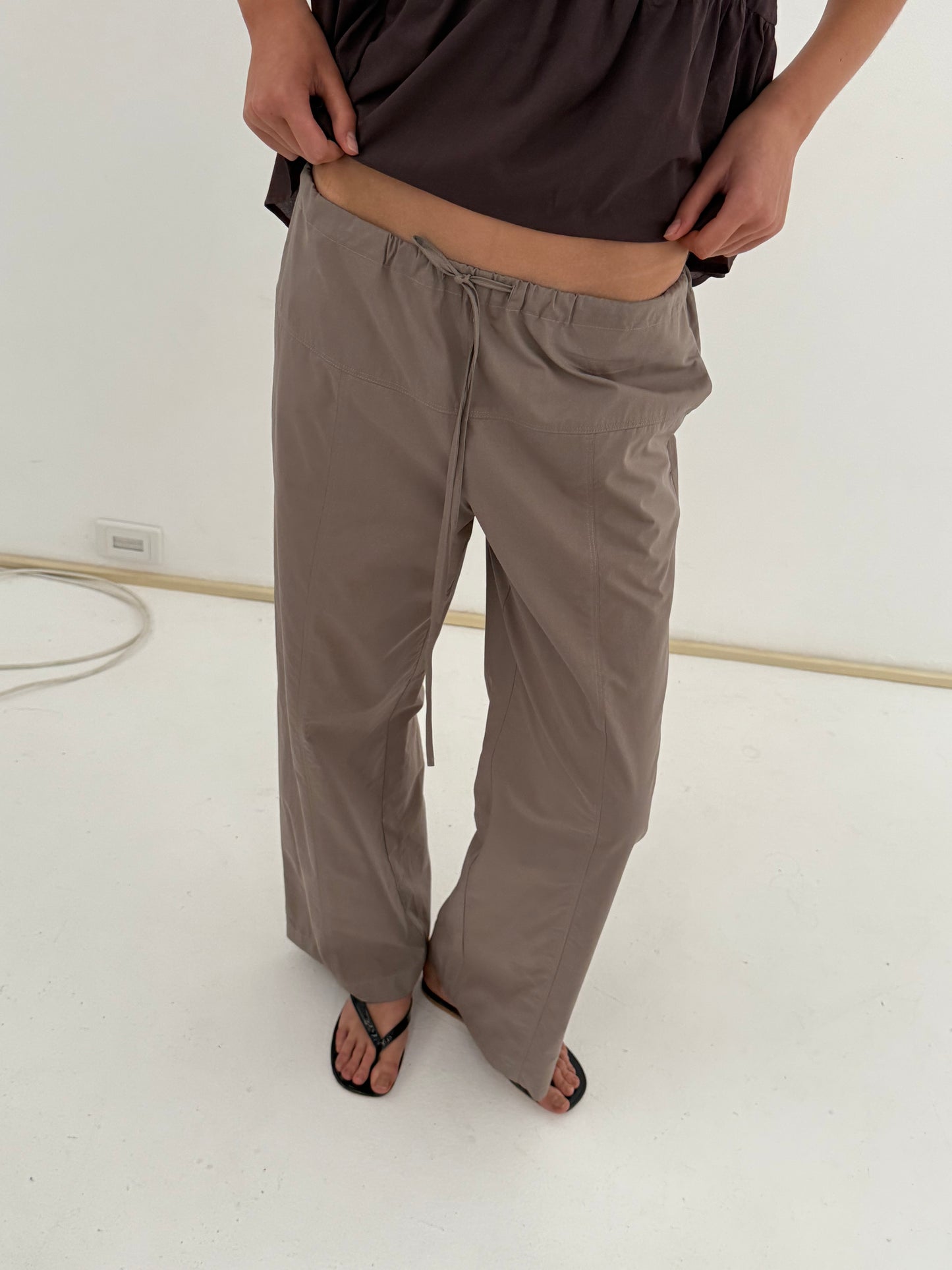 Person wearing Deiji Studios’ Seam Pant in Light Khaki, styled with black flip-flops and a dark top. Hands rest near waistband on a minimalist white background; face is not visible.
