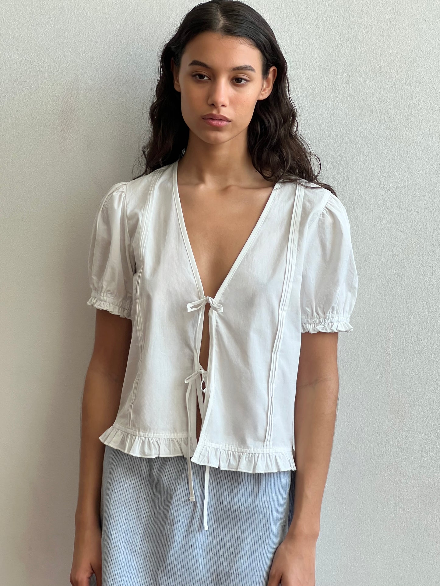 A woman with long dark hair wears the Deiji Studios Pintuck Top in white—an organic cotton poplin blouse with a deep V-neck, tie closure, puffed sleeves, and ruffled hem—paired with a light blue skirt against a plain light wall.