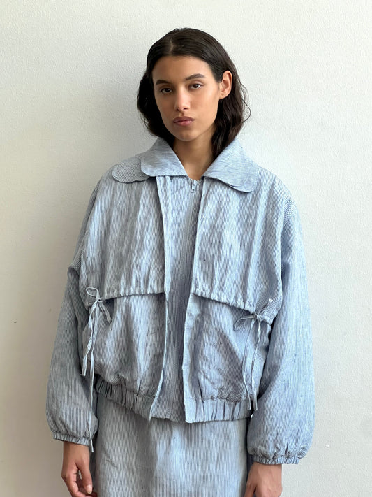 A woman with long dark hair stands against a white wall, wearing the Double Layered Jacket - Blue Stripe, a loose-fitting piece with large front pockets and matching pants. She keeps a neutral expression with her hands at her sides.