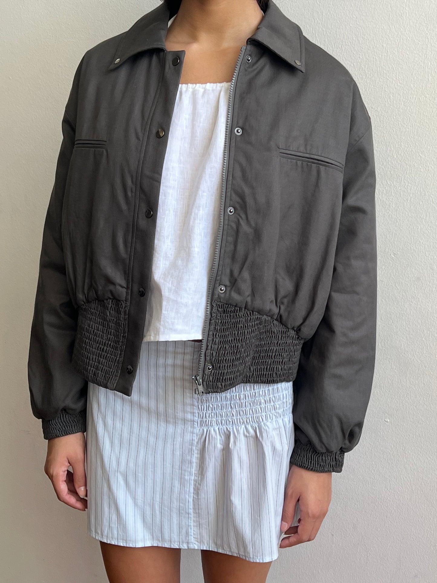 A person wears the Shirring Jacket - Slate by Deiji Studios, featuring elastic cuffs and hem, over a white sleeveless top and a white skirt with blue stripes, standing against a light-colored wall.