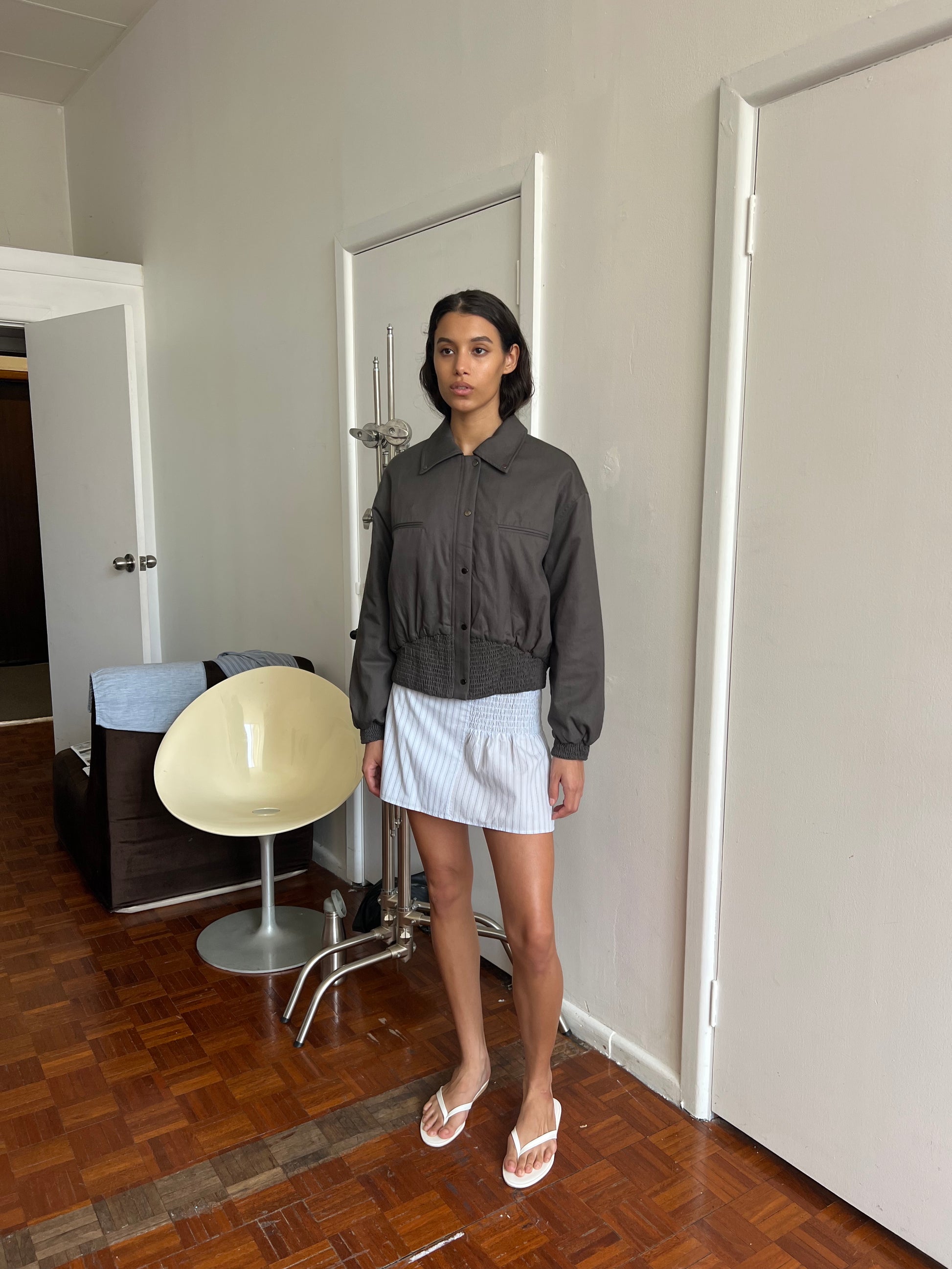 A woman indoors on a wooden floor wears a dark gray jacket, white flip-flops, and the Deiji Studios Shirring Skirt - Mineral Stripe. A modern white chair, metal coat rack, and closed white doors are seen in the background.