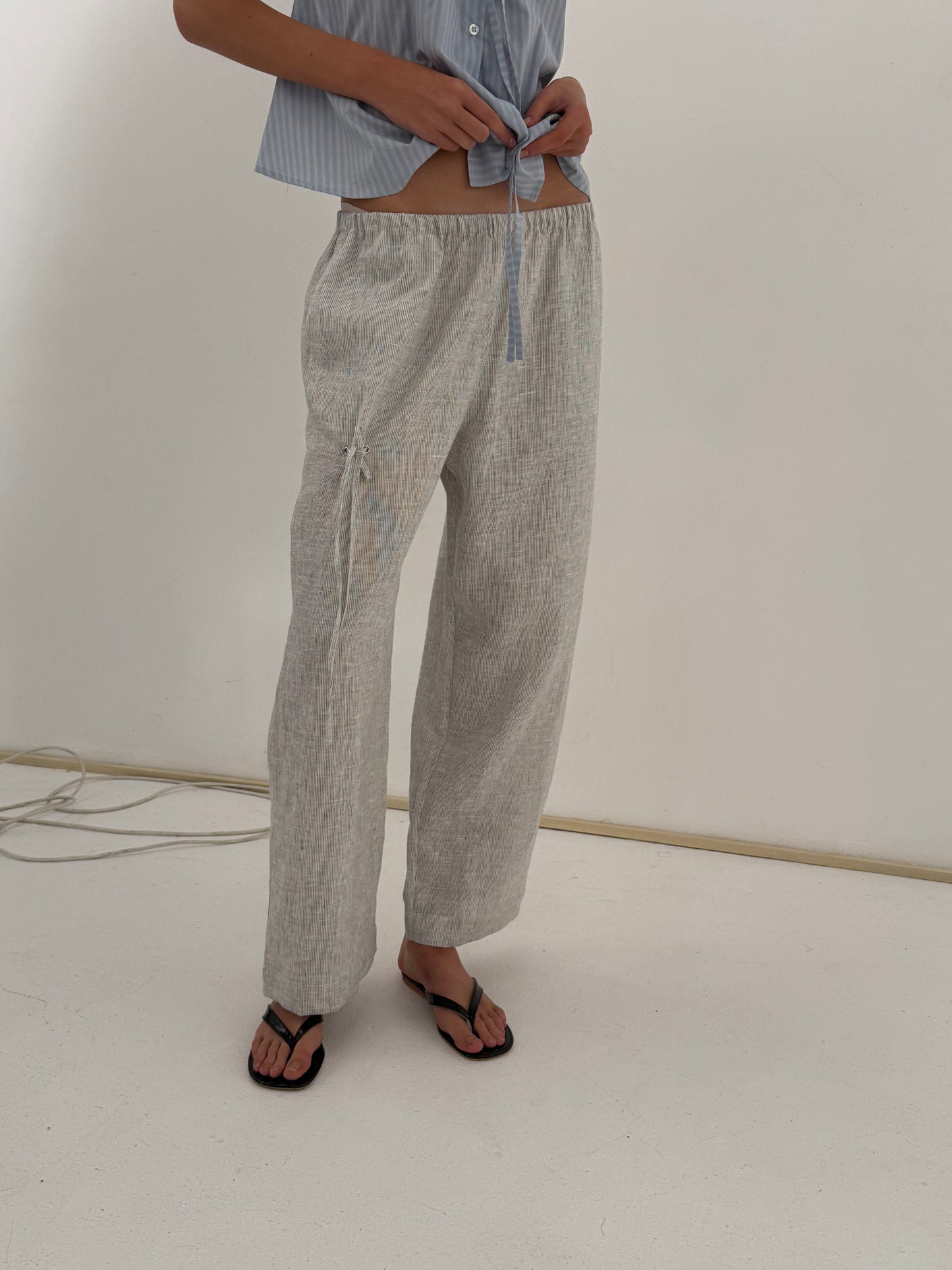 A person wears the Eyelet Pant - Fine Stripe, ankle length and relaxed, with a light blue shirt tied at the waist and black sandals, standing on a white floor against a plain wall—Deiji Studios-inspired style.