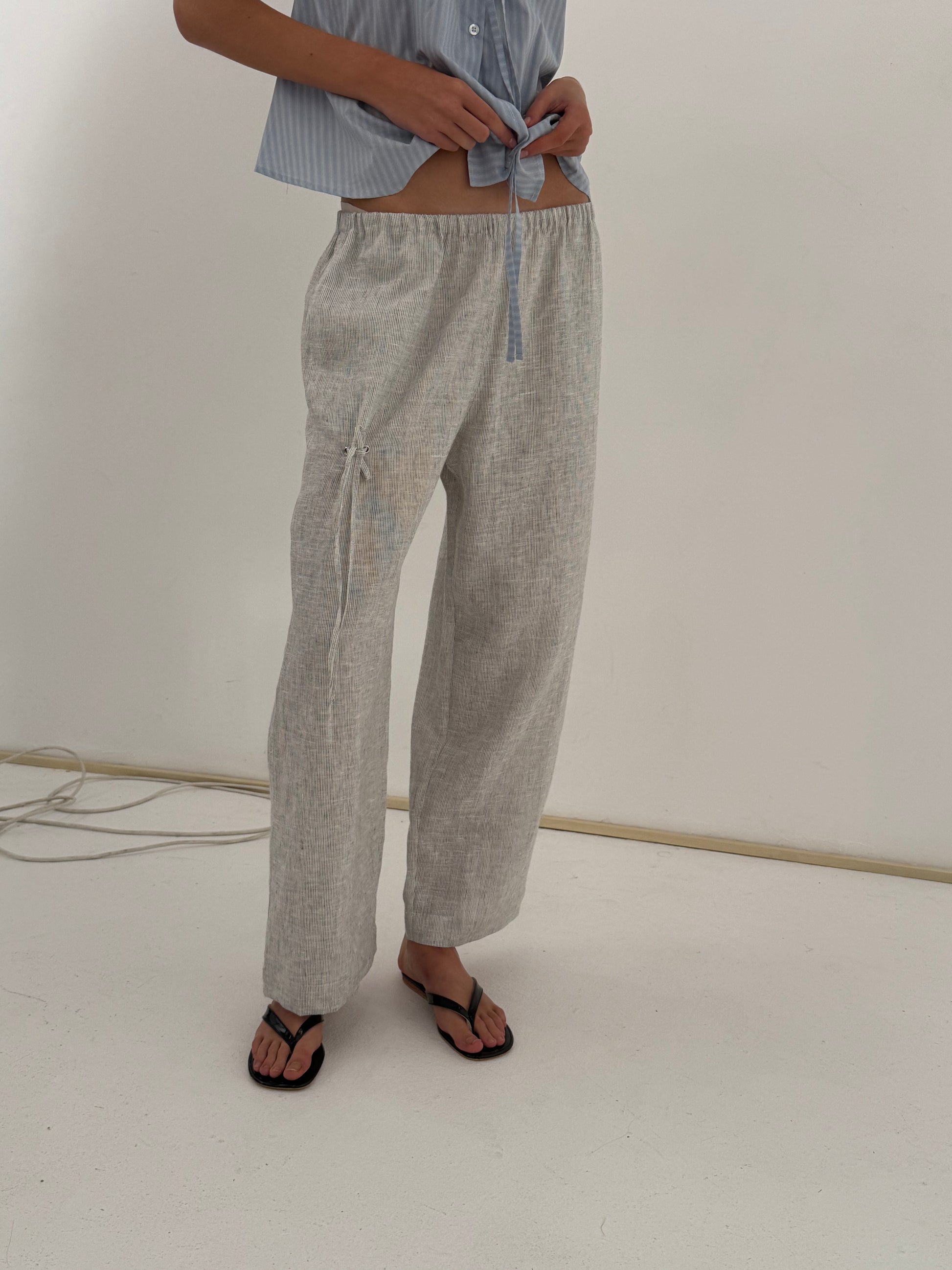A person wears the Eyelet Pant - Fine Stripe, ankle length and relaxed, with a light blue shirt tied at the waist and black sandals, standing on a white floor against a plain wall—Deiji Studios-inspired style.
