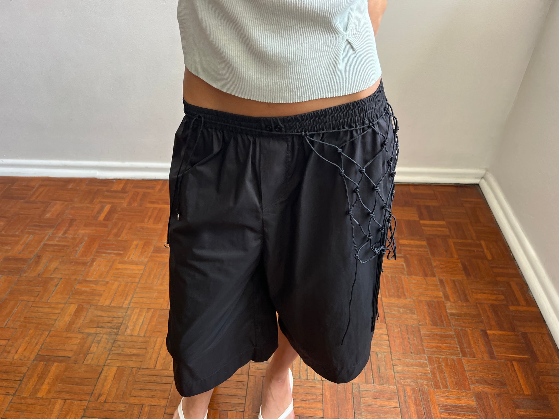 A person wearing the Long Lounger - Black stands on a wooden parquet floor against a plain white wall, embodying Deiji Studios’ relaxed elegance.