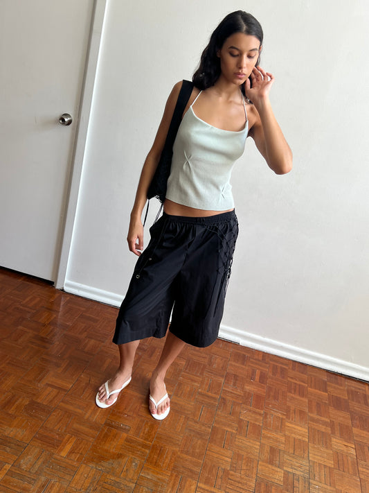 A woman stands indoors on a wooden floor, wearing a light blue camisole, the Long Lounger - Black from Deiji Studios, white flip-flops, and carrying a black shoulder bag. She touches her ear and gazes down thoughtfully.
