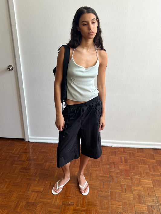 A woman with long dark hair stands indoors on a wooden floor, wearing an organic cotton light blue tank top, black knee-length shorts called the Long Lounger - Black, white flip-flops, and a black backpack from Deiji Studios, looking off to the side.