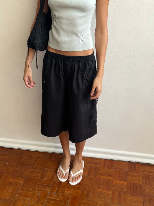 A woman stands indoors on a wooden floor near a white wall, wearing the Long Lounger - Black with textured side detail, a light blue sleeveless top, white flip-flops, and carrying a black woven bag from Deiji Studios.