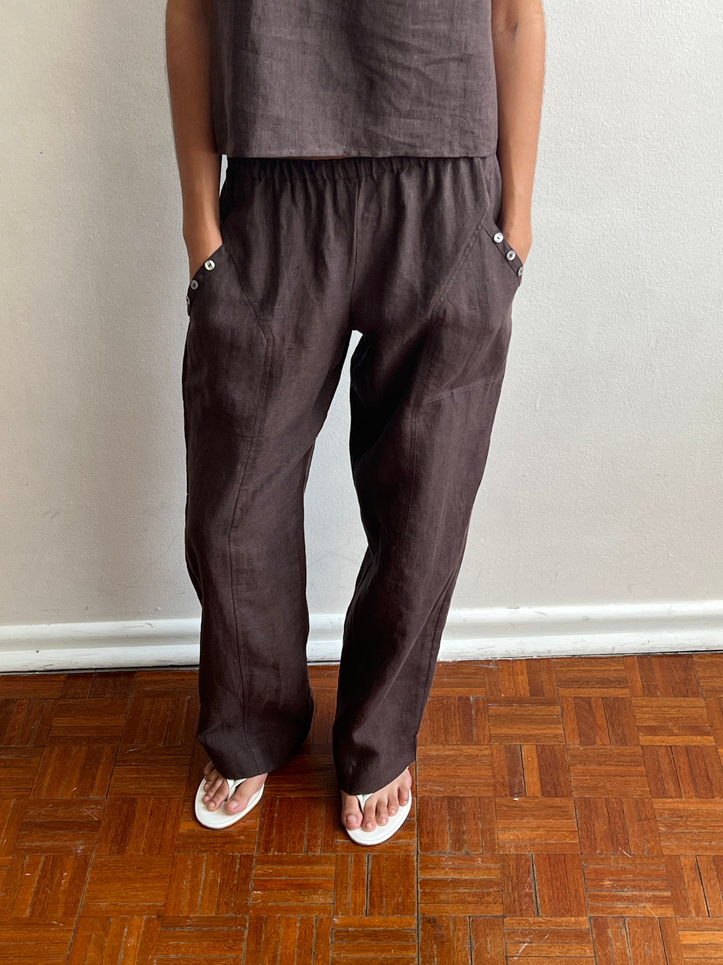 A person stands on a wooden floor against a light wall, wearing the Angle Pocket Pant - Bark by Deiji Studios, styled with hands in pockets, a matching top, and white flip-flops.