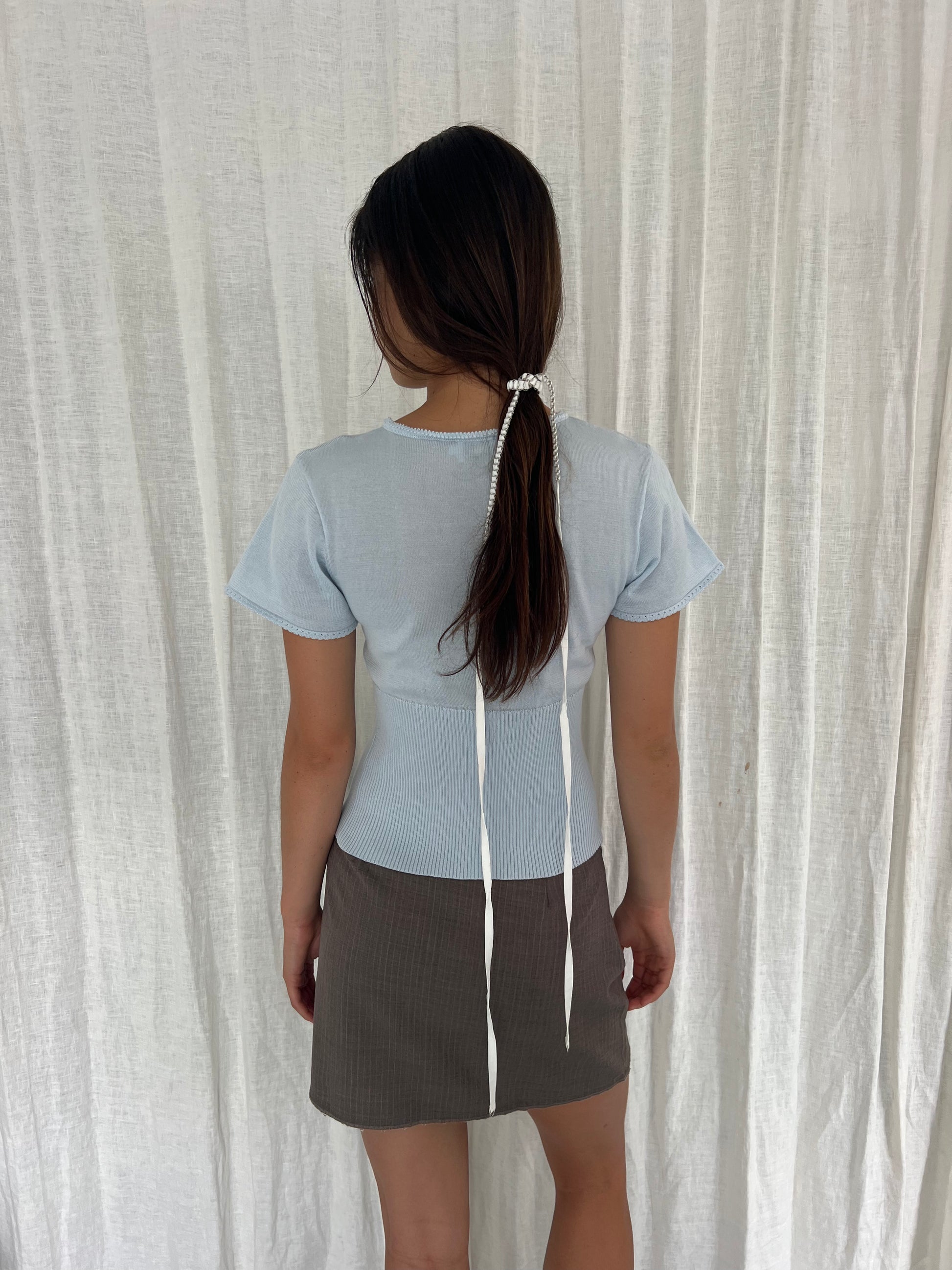 A woman faces away, wearing the Trim Knit Tee - Ice Blue from Deiji Studios and a brown skirt. Her long hair is tied back with a white beaded accessory and two white ribbons, standing before a white curtain.
