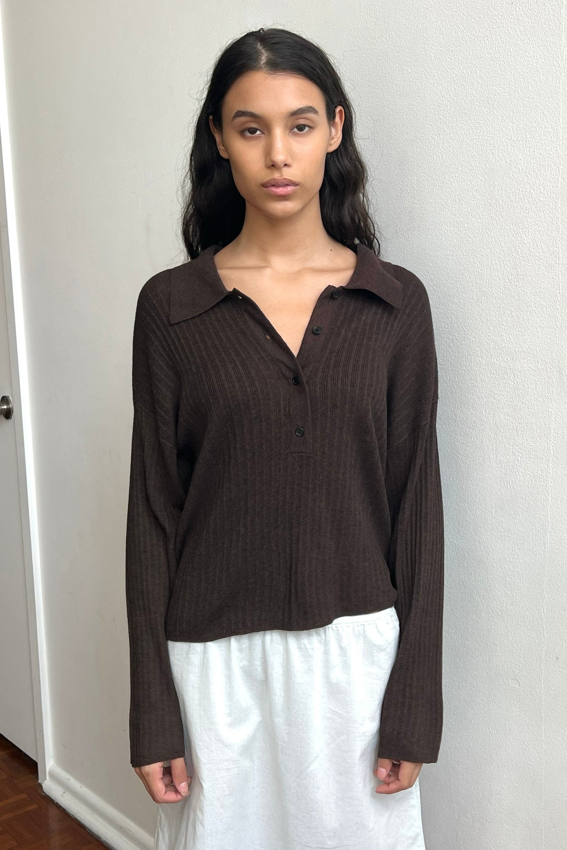 A woman with long dark hair stands indoors against a white wall, wearing the Polo Knit - Dark Almond by Deiji Studios with buttons and a white skirt, looking directly at the camera with a neutral expression.