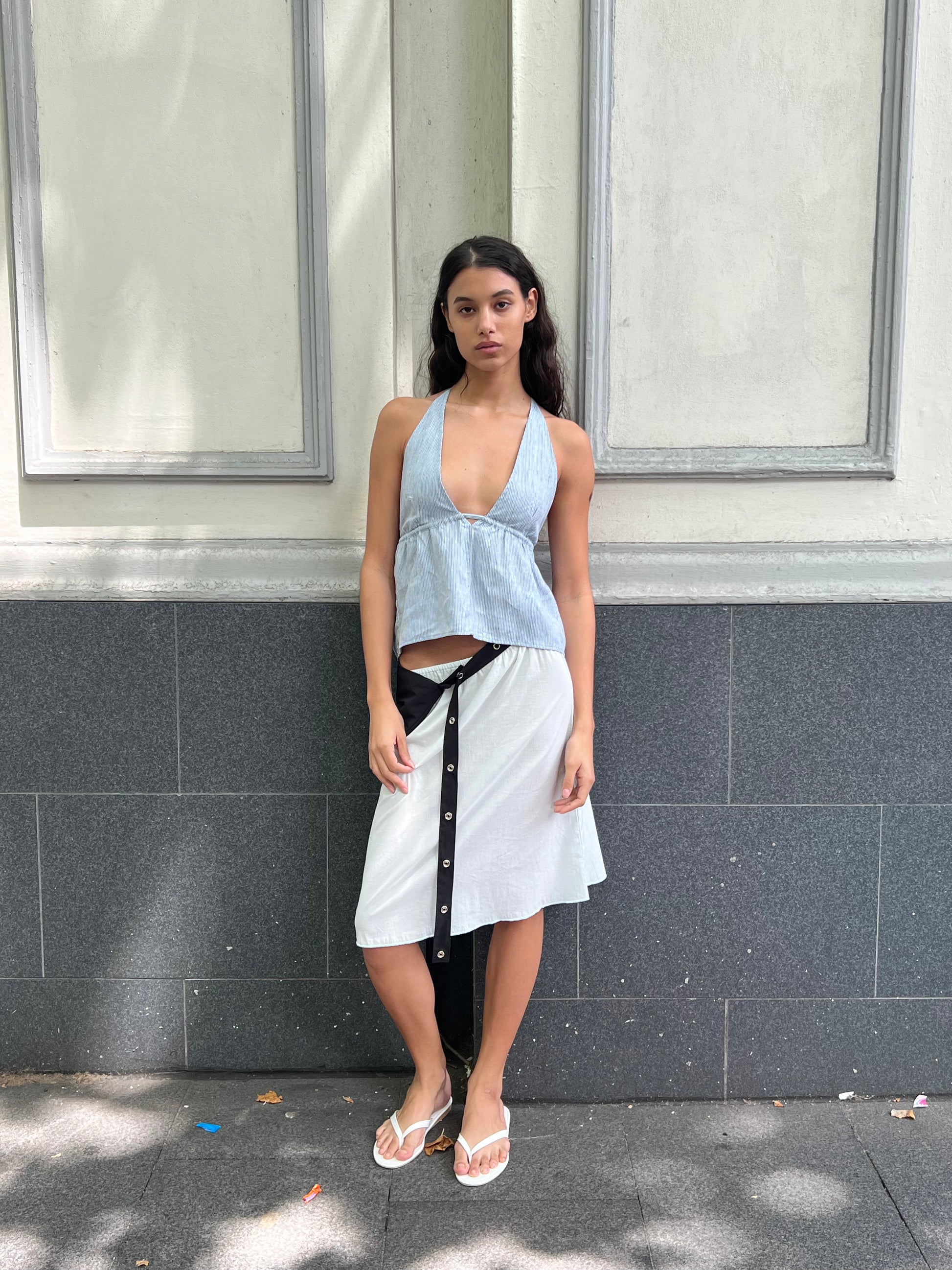 A woman with long dark hair stands against a gray and white wall, wearing the Linen Button Halter - Blue Stripe from Deiji Studios, a white wrap skirt with black buttons, and white flip-flops. She gazes neutrally at the camera.