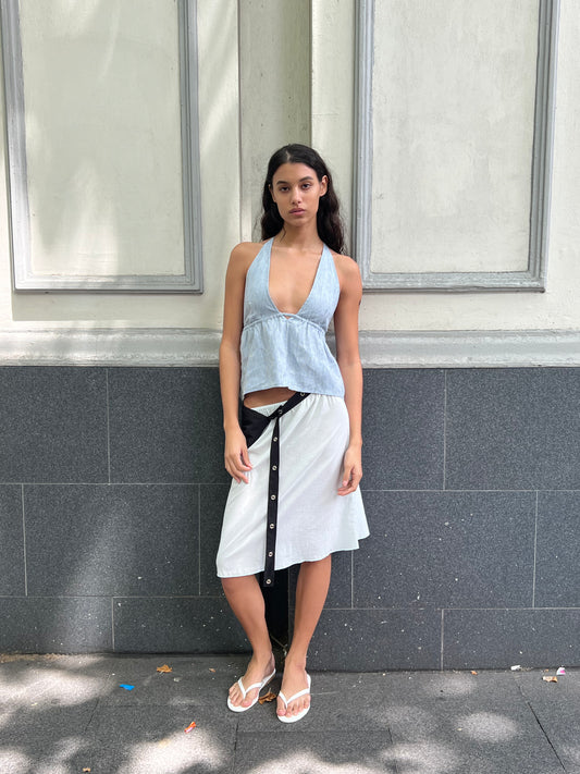 A woman with long dark hair stands against a gray and white wall, wearing the Linen Button Halter - Blue Stripe from Deiji Studios, a white wrap skirt with black buttons, and white flip-flops. She gazes neutrally at the camera.