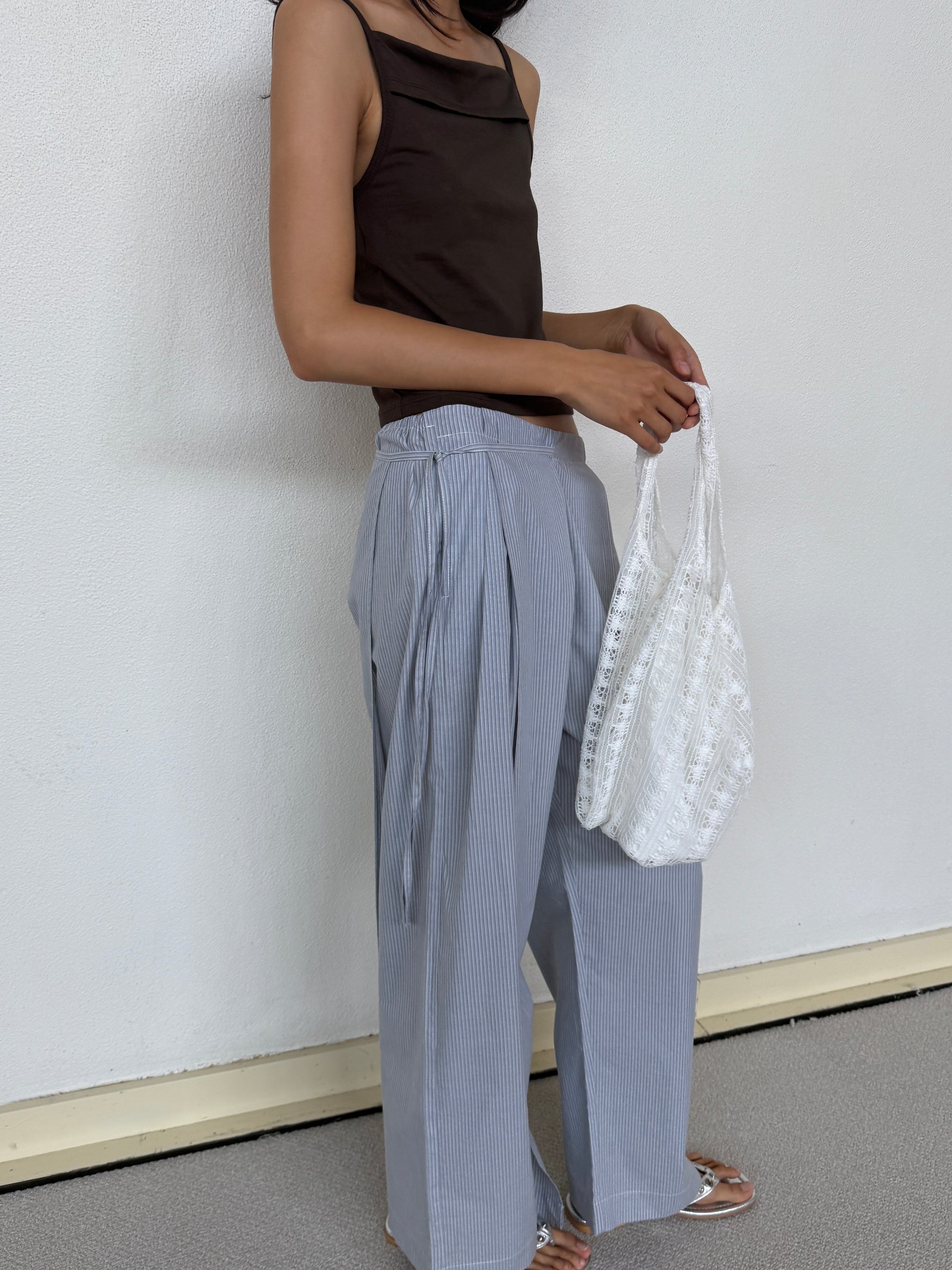 A person stands against a white wall, wearing a sleeveless brown top, loose light blue striped pants, sandals, and holding the Thread Dust Bag in Off White. Their face is partially out of frame.