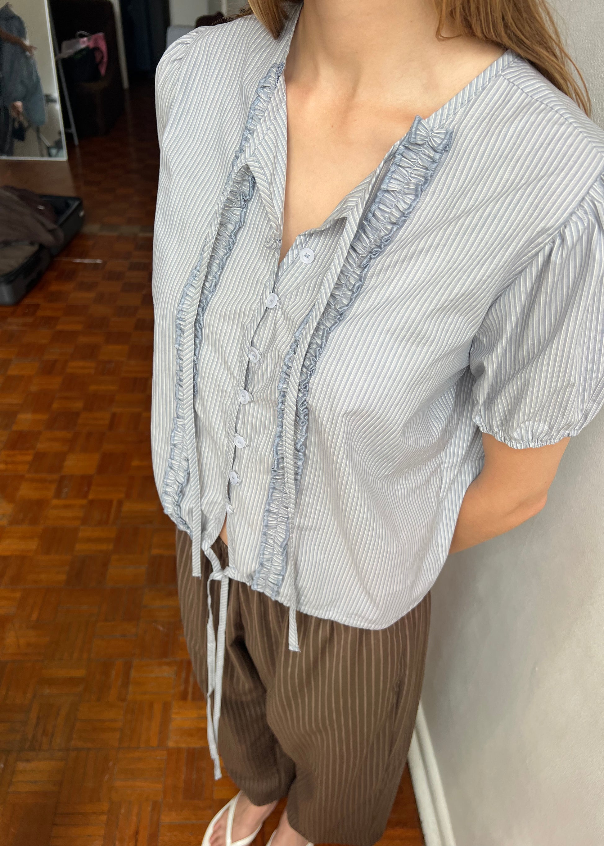 Indoors, a person wears the Double Frill Top - Sky Stripe with ruffles and buttons, styled with brown pinstriped pants and white sandals. Their arms are placed behind their back.