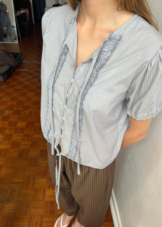Indoors, a person wears the Double Frill Top - Sky Stripe with ruffles and buttons, styled with brown pinstriped pants and white sandals. Their arms are placed behind their back.