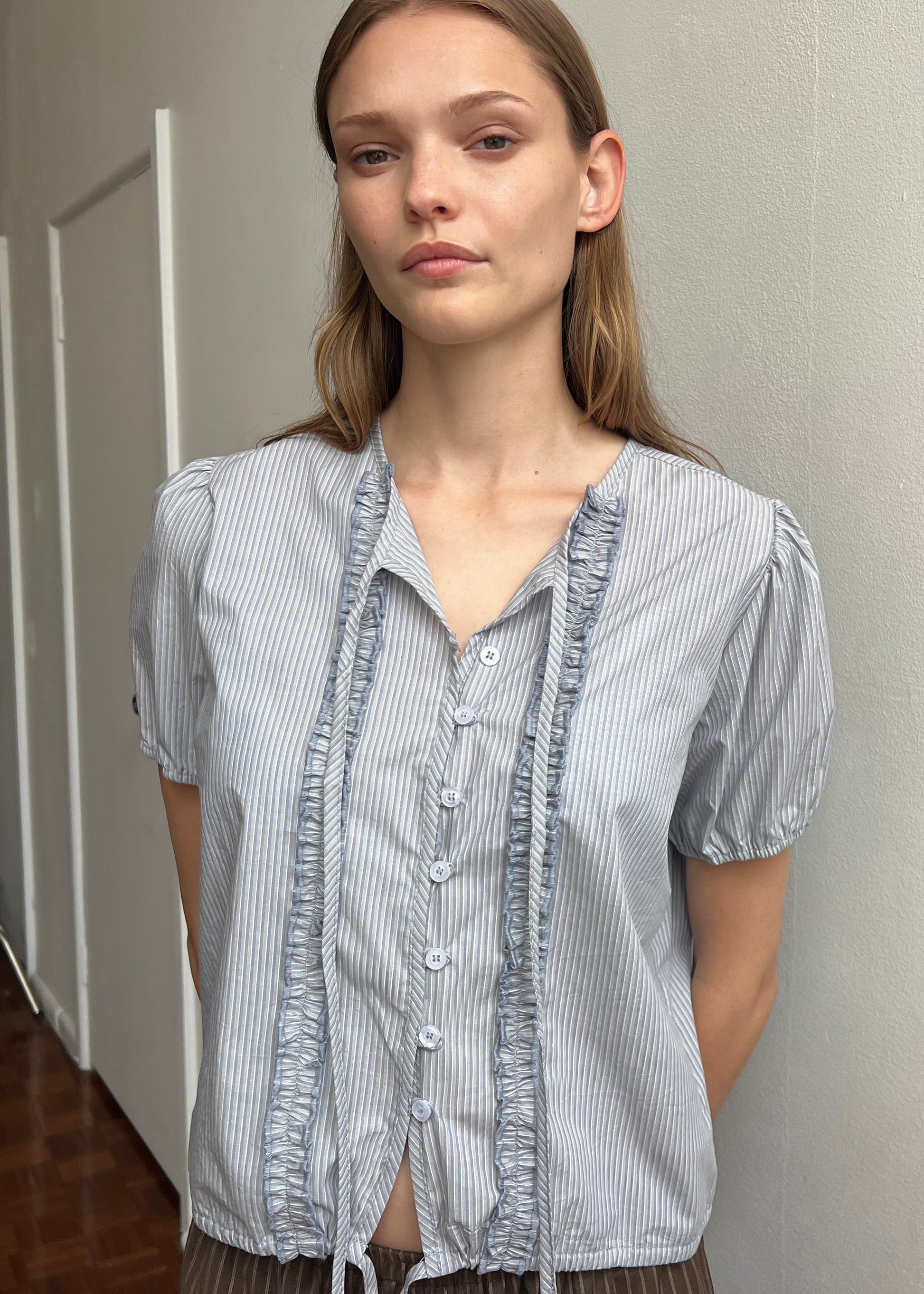 A woman with long light brown hair stands against a light wall, wearing the Double Frill Top - Sky Stripe by Deiji Studios, made from organic cotton poplin with blue and white stripes and ruffle details. She looks ahead with a neutral expression.