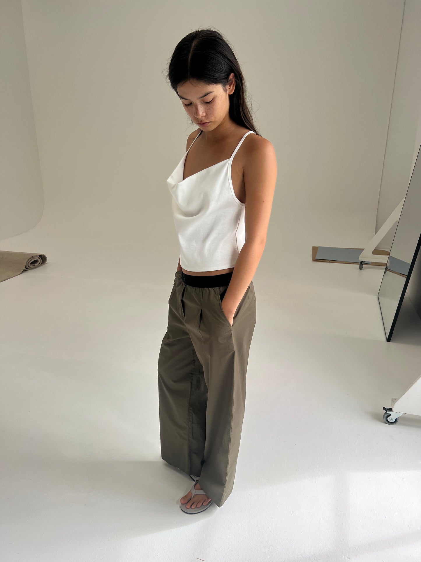 A woman stands indoors on a white floor, looking down. She wears a Deiji Studios white sleeveless top and the Contrast Waist Pant - Olive, paired with sandals. The minimalistic setting has neutral walls and subtle studio props.