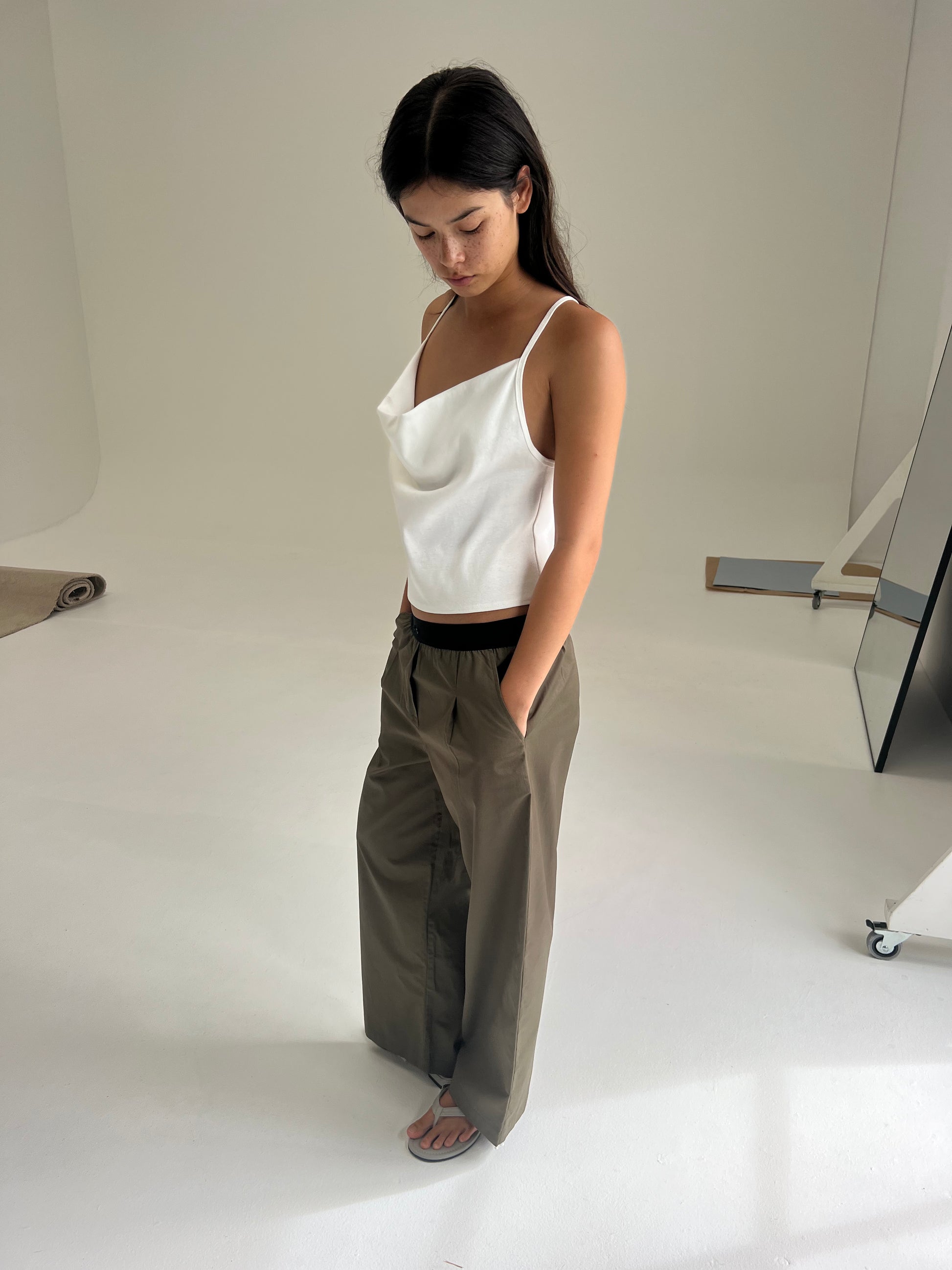 A woman stands indoors on a white floor, looking down. She wears a Deiji Studios white sleeveless top and the Contrast Waist Pant - Olive, paired with sandals. The minimalistic setting has neutral walls and subtle studio props.