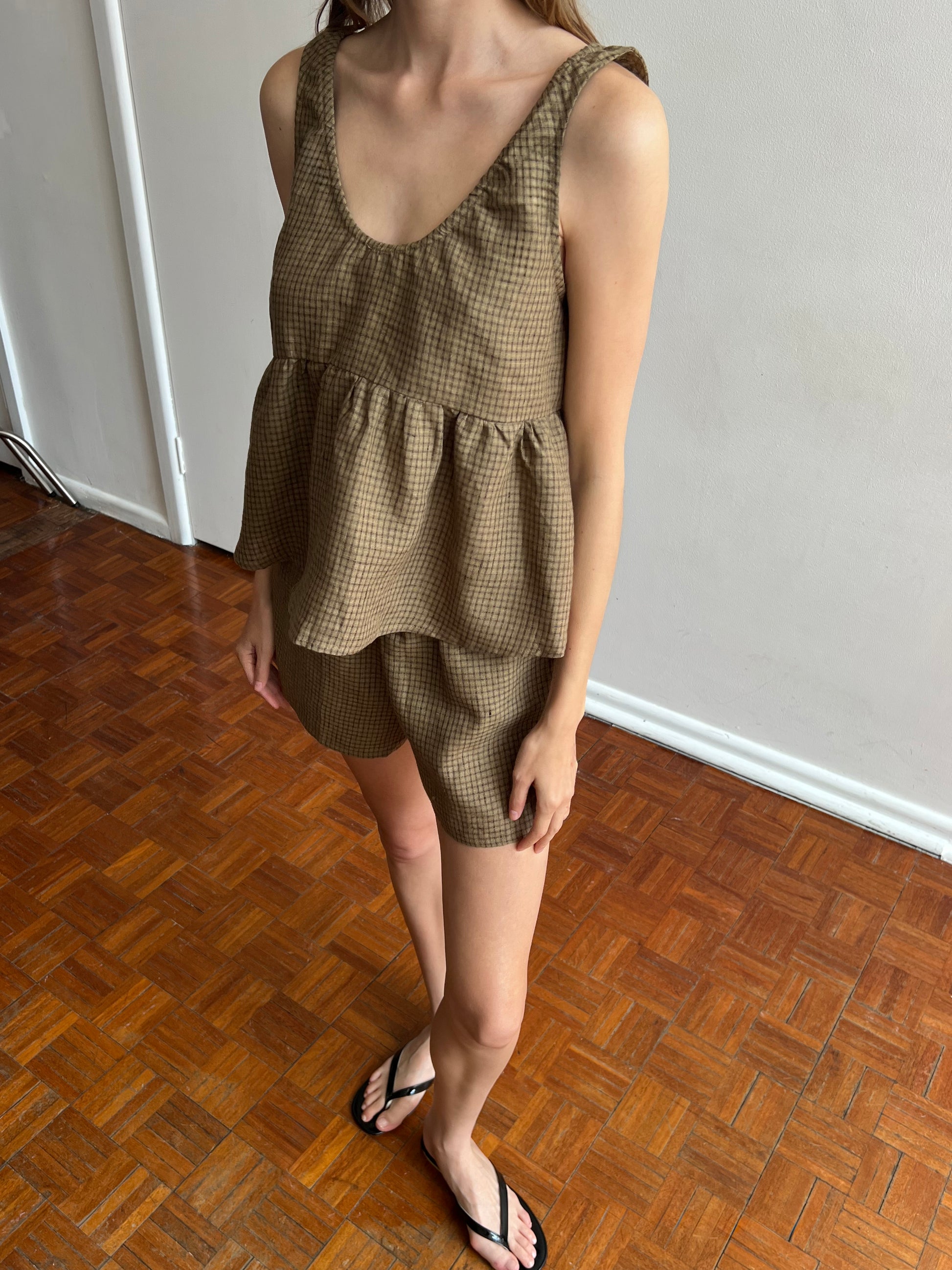 A woman stands indoors on a parquet floor wearing the Gathered Linen Set - Pine Check from Deiji Studios, featuring a sleeveless peplum top and matching shorts with black flip-flops. Her head is partially out of frame.