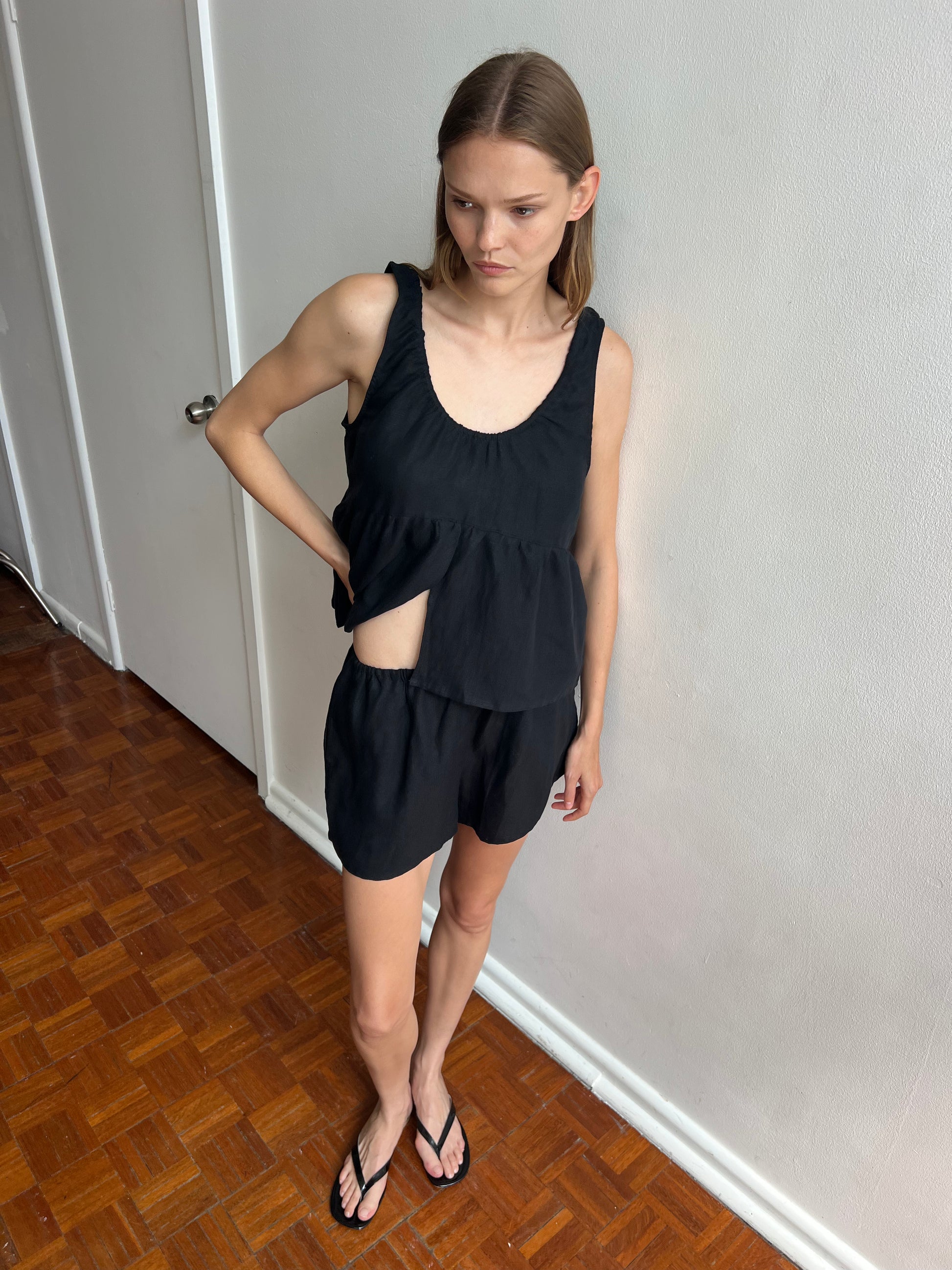 A woman stands indoors on a wooden floor wearing Deiji Studios’ Gathered Linen Set - Black, featuring a sleeveless black top with cutout detail and matching shorts. She looks down and to the side, with one hand on her waist.