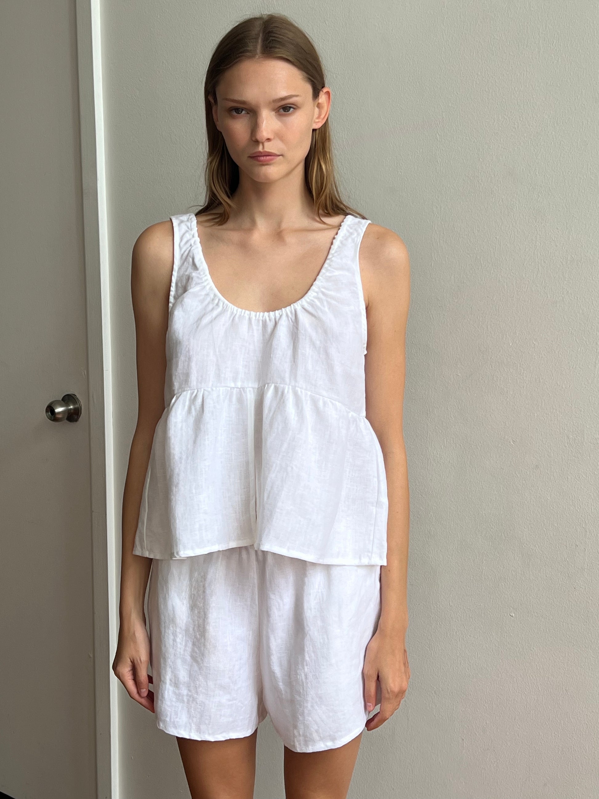 A woman with straight, light brown hair and a neutral expression stands indoors against a plain light wall, wearing the Gathered Linen Set - White: a sleeveless, loose-fitting linen top with peplum hem and matching shorts.