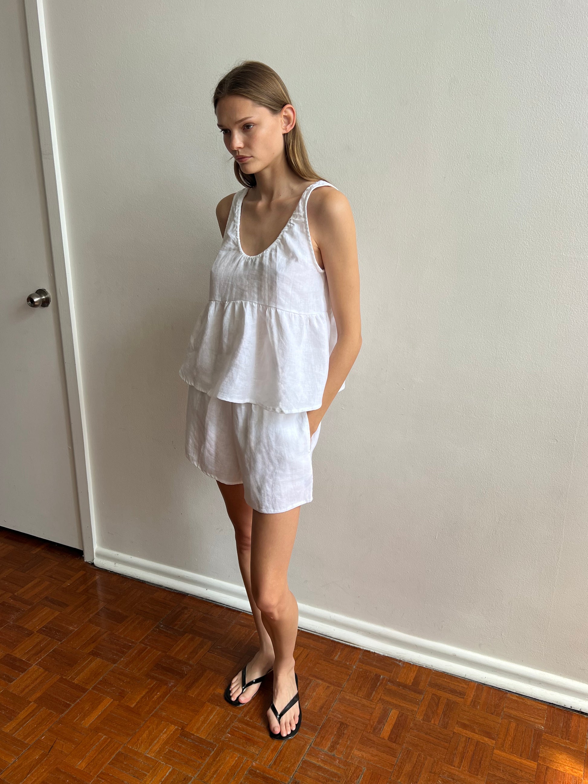 A woman stands indoors on a wooden floor, wearing the Gathered Linen Set - White with black flip-flops, beside a light-colored wall and a closed door.